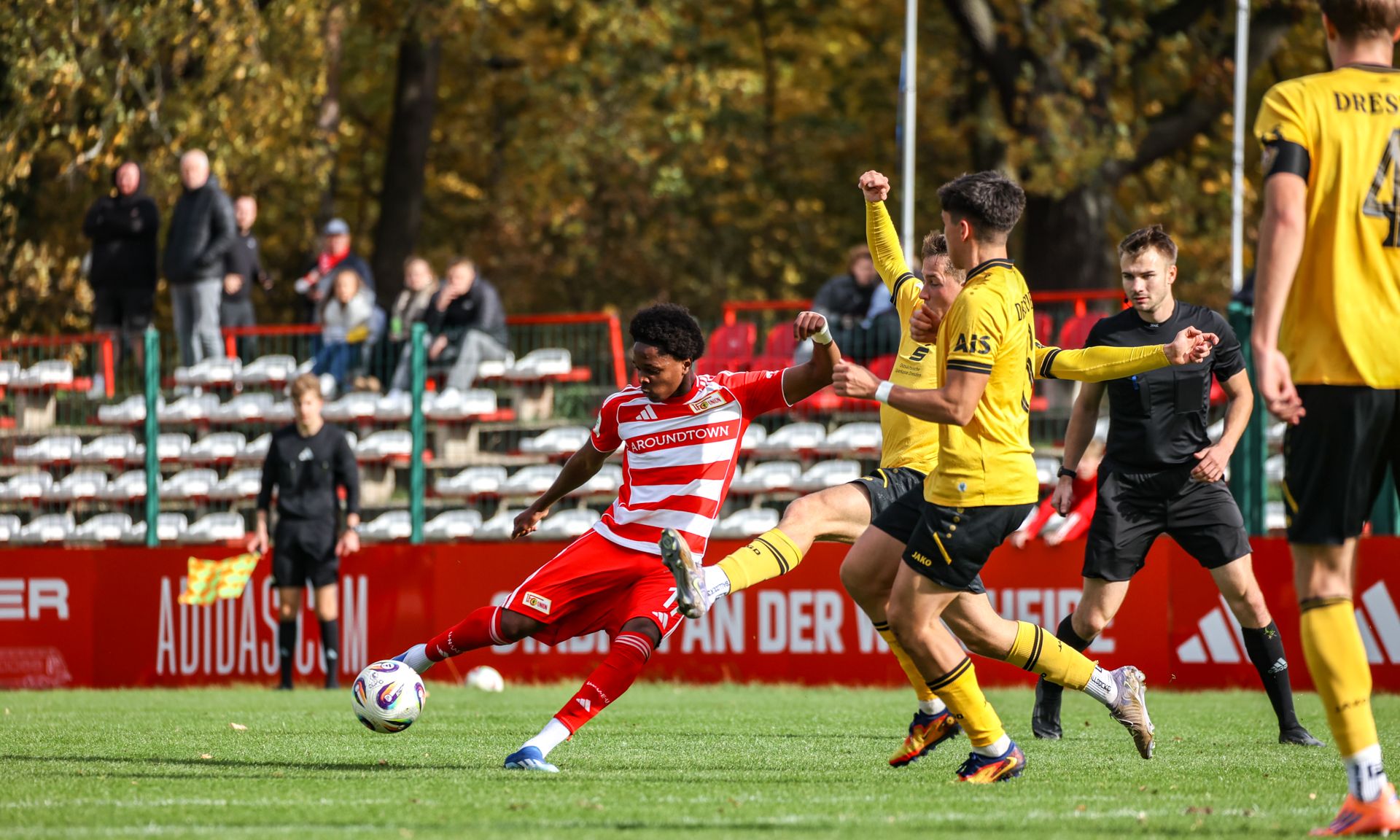 Ein Fußballspieler in rotem Trikot fällt im Spiel, während ein gegnerischer Spieler in gelbem Trikot ihn herausfordert.