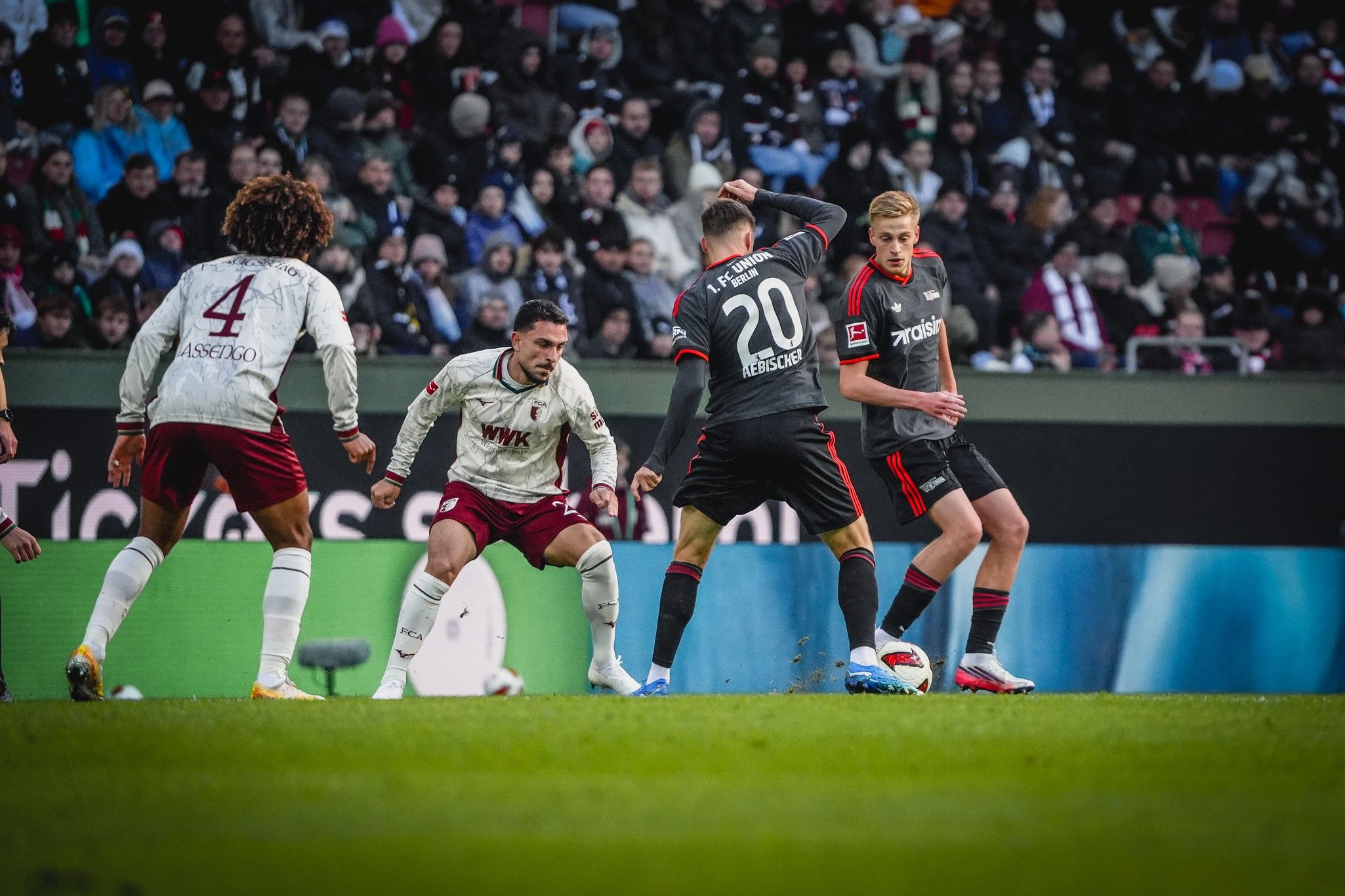 Ein Fußballspiel mit mehreren Spielern auf dem Rasen, einer in der Mitte dribbelt, während andere am Ball verteidigen.