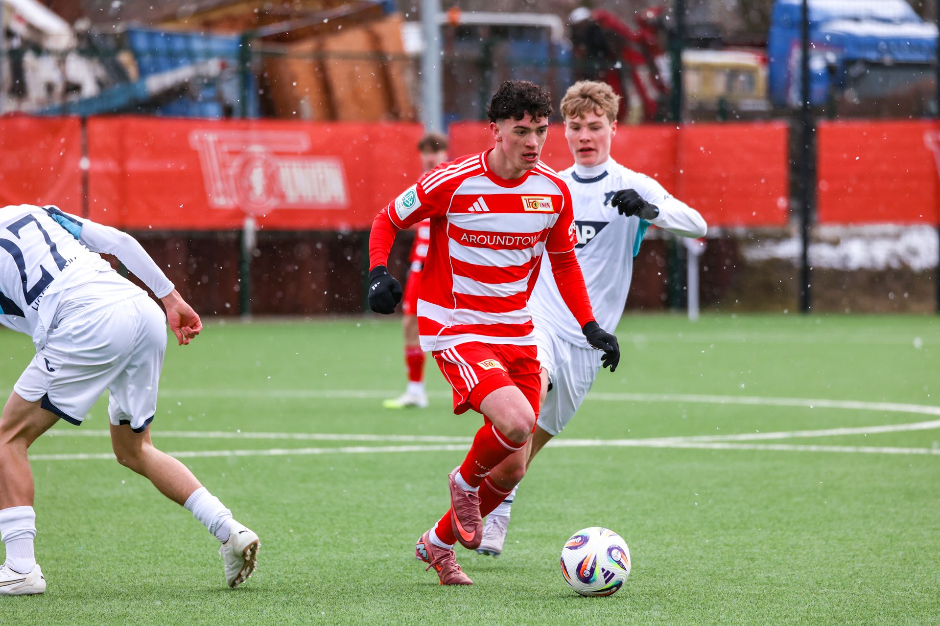 Junger Fußballer in rotem Trikot dribbelt den Ball auf einem Sportplatz, während zwei Spieler ihn verfolgen. Schnee fällt.