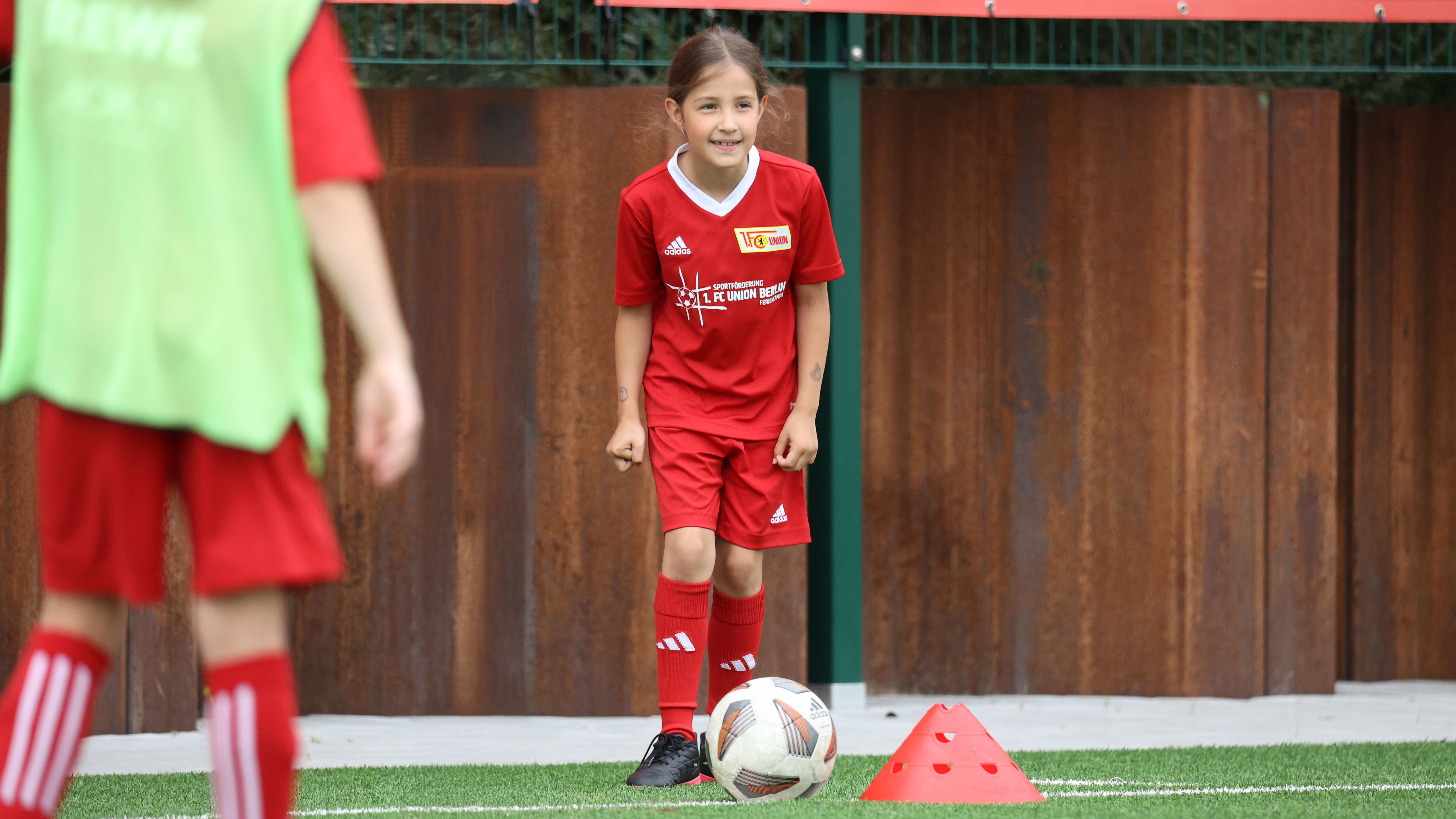 Ein Mädchen in rotem Fußballtrikot steht auf dem Spielfeld und lächelt, während sie auf einen Ball und Hütchen schaut.