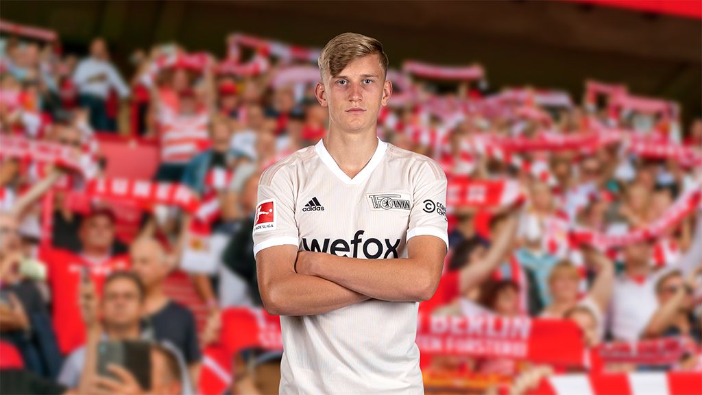 Ein junger Fußballspieler in einem weißen Trikot steht mit verschränkten Armen vor einer jubelnden Menschenmenge im Stadion.