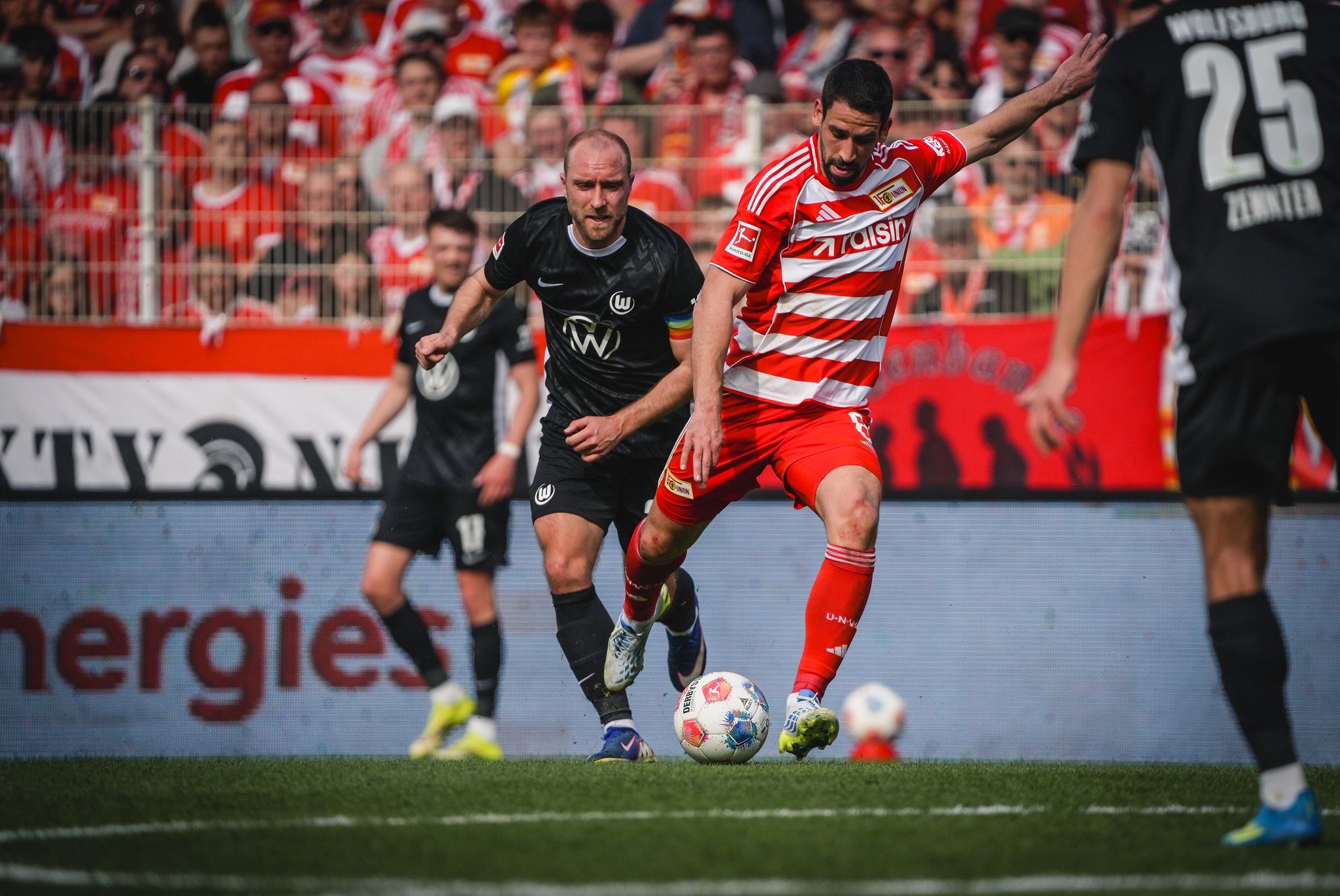 Fußballspieler in rot-weißen Trikots bewegt sich mit dem Ball, während ein Spieler in schwarz versucht, ihn zu stoppen.
