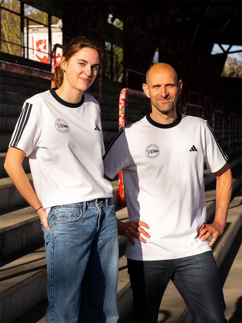 Zwei Personen stehen auf einer Tribüne, tragen passende weiße T-Shirts mit schwarzen Streifen und Jeans.