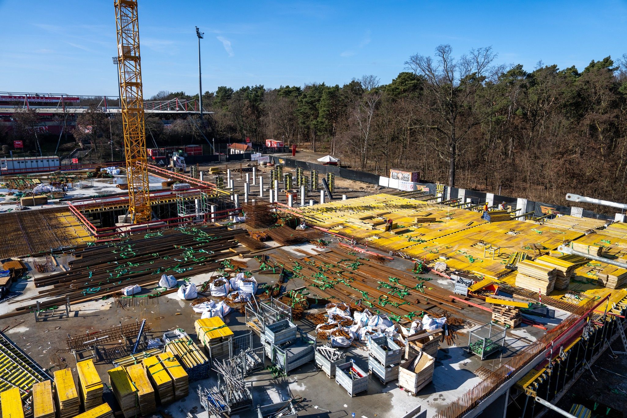 Baustelle mit gelben Materialien, Baustellenkran und unvollendeten Strukturen, umgeben von Bäumen und einem blauen Himmel.
