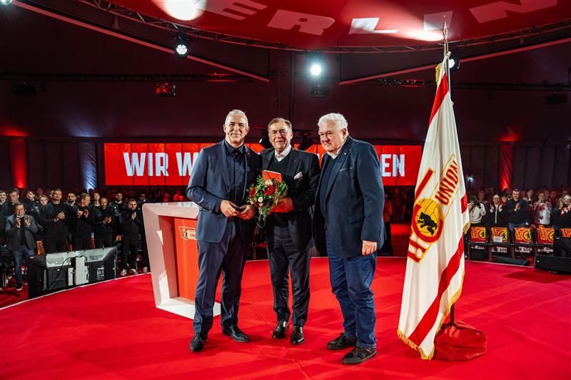 Drei Männer stehen auf einer Bühne, umgeben von Publikum, mit einem Blumenstrauß und einer Flagge im Hintergrund.