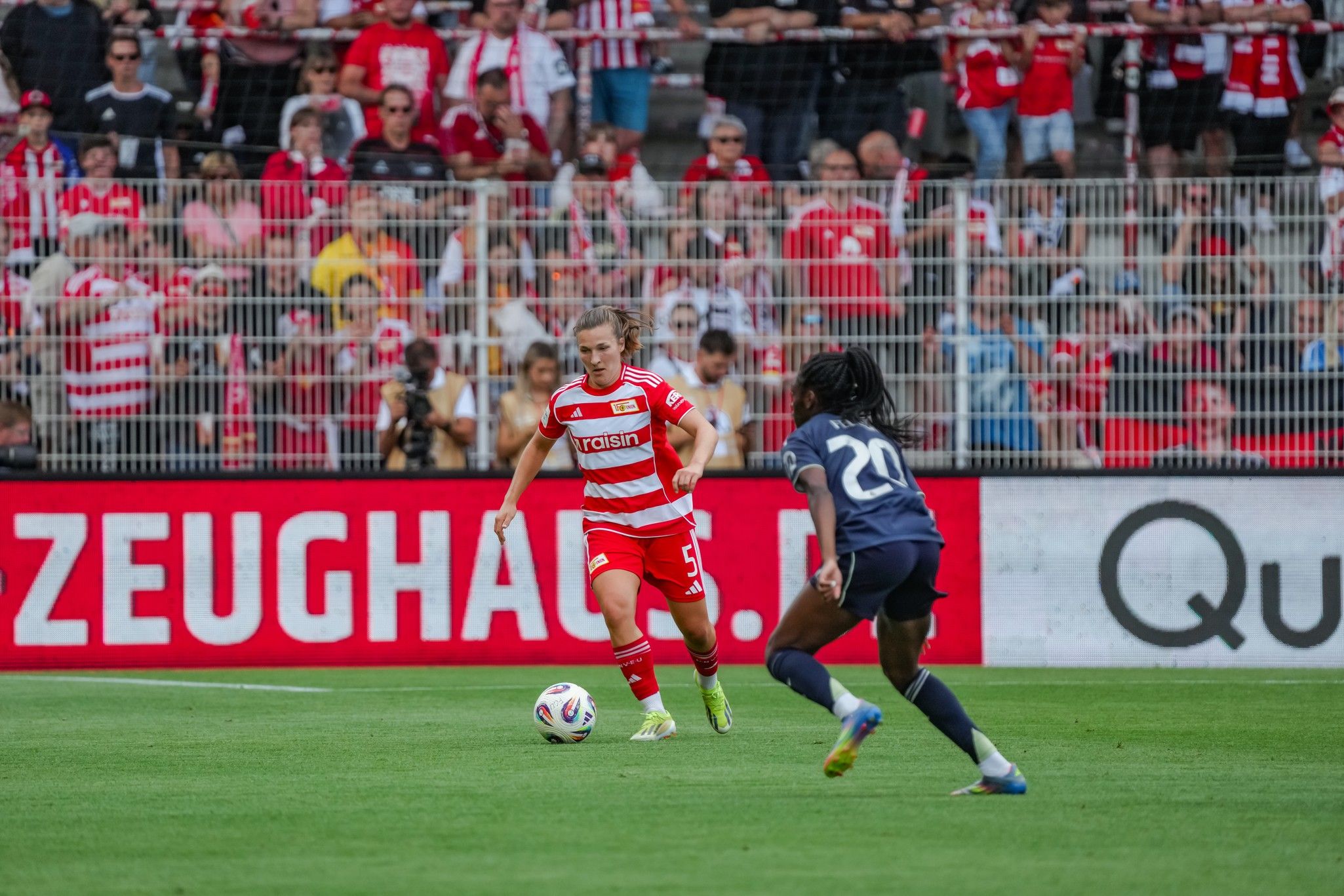 Eine Fußballspielszene mit einem Spieler in rot-weißer Kleidung, der den Ball dribbelt, während ein gegnerischer Spieler ihn verfolgt.