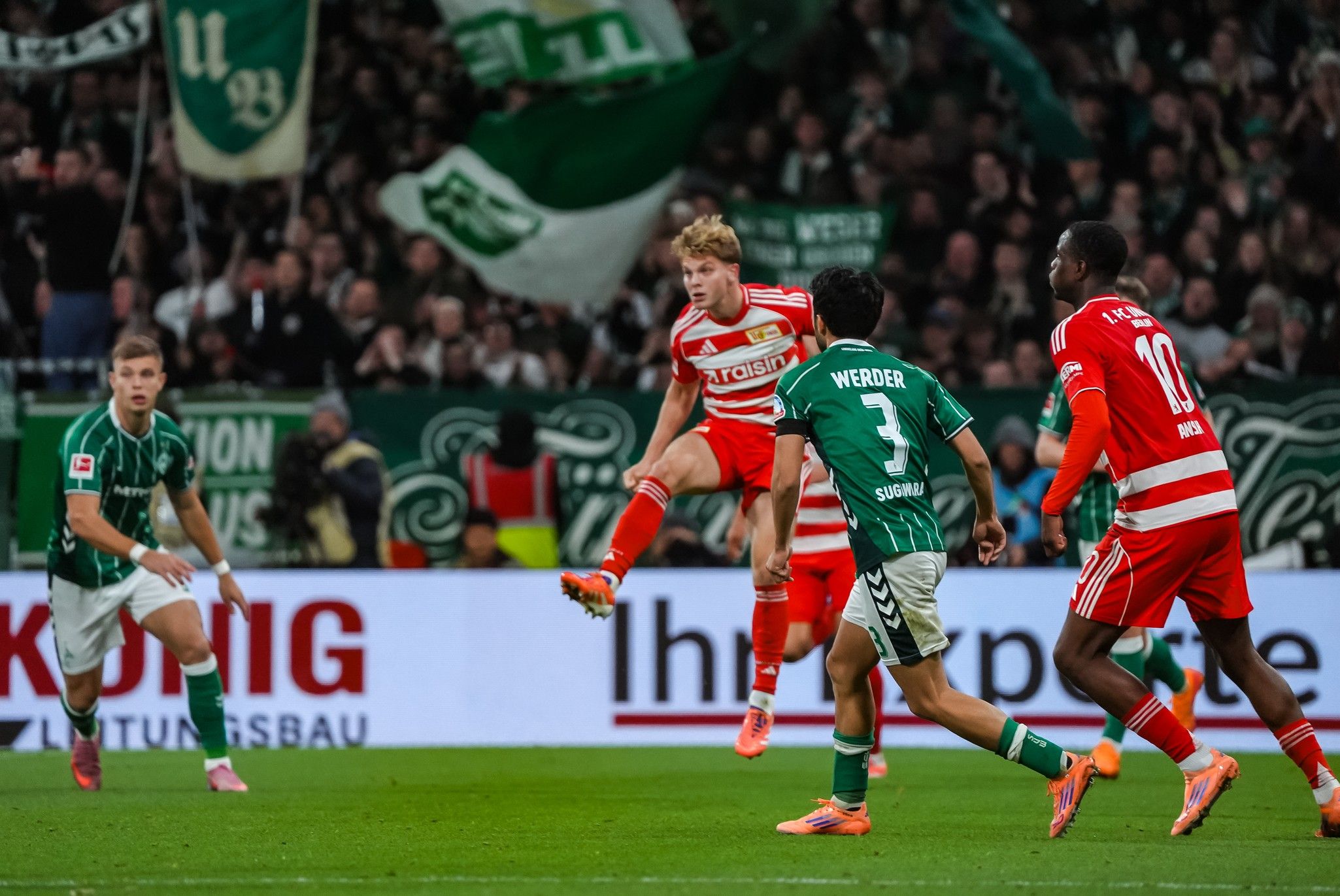 Ein Fußballspieler springt zum Schuss, während andere Spieler und Fans im Hintergrund zu sehen sind.