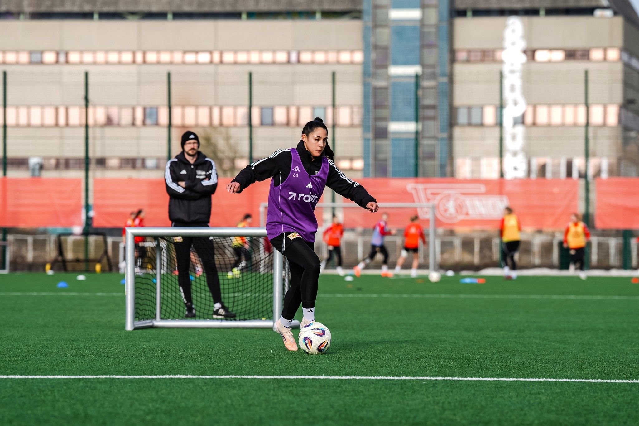 Eine Fußballspielerin in lila Trikot schießt den Ball auf ein kleines Tor während des Trainings auf einem Kunstrasenplatz.