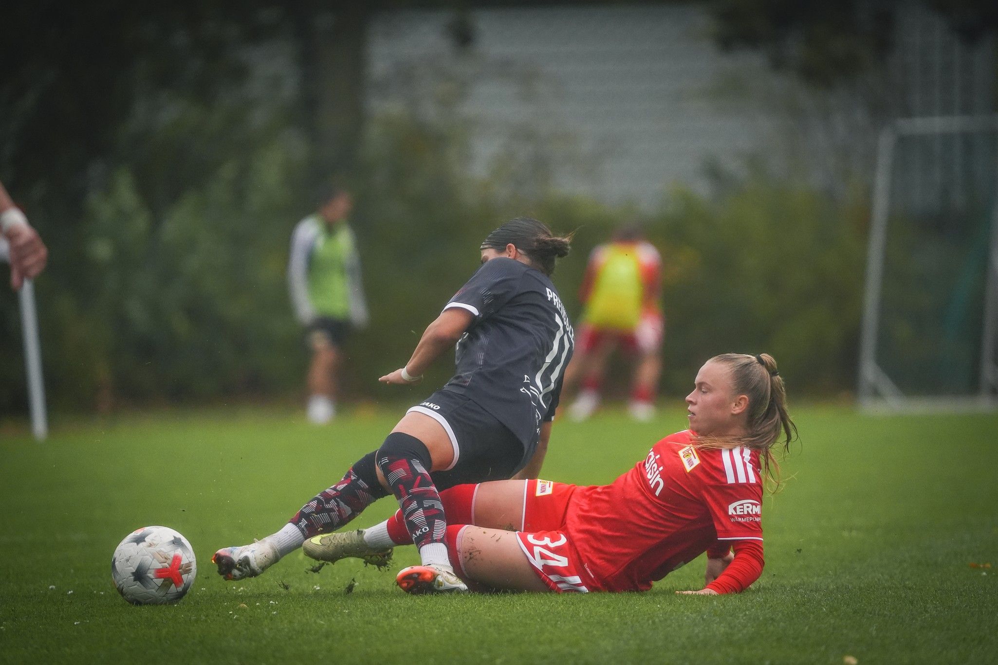 Zwei Fußballspielerinnen im Rasen, eine fällt zu Boden, während die andere sie herausfordert. Beide tragen Sportkleidung und Schienbeinschützer.