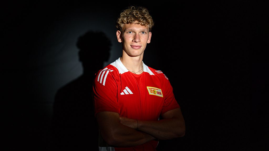 Ein junger Mann mit lockigem blondem Haar steht in einem roten Fußballtrikot vor einem dunklen Hintergrund.