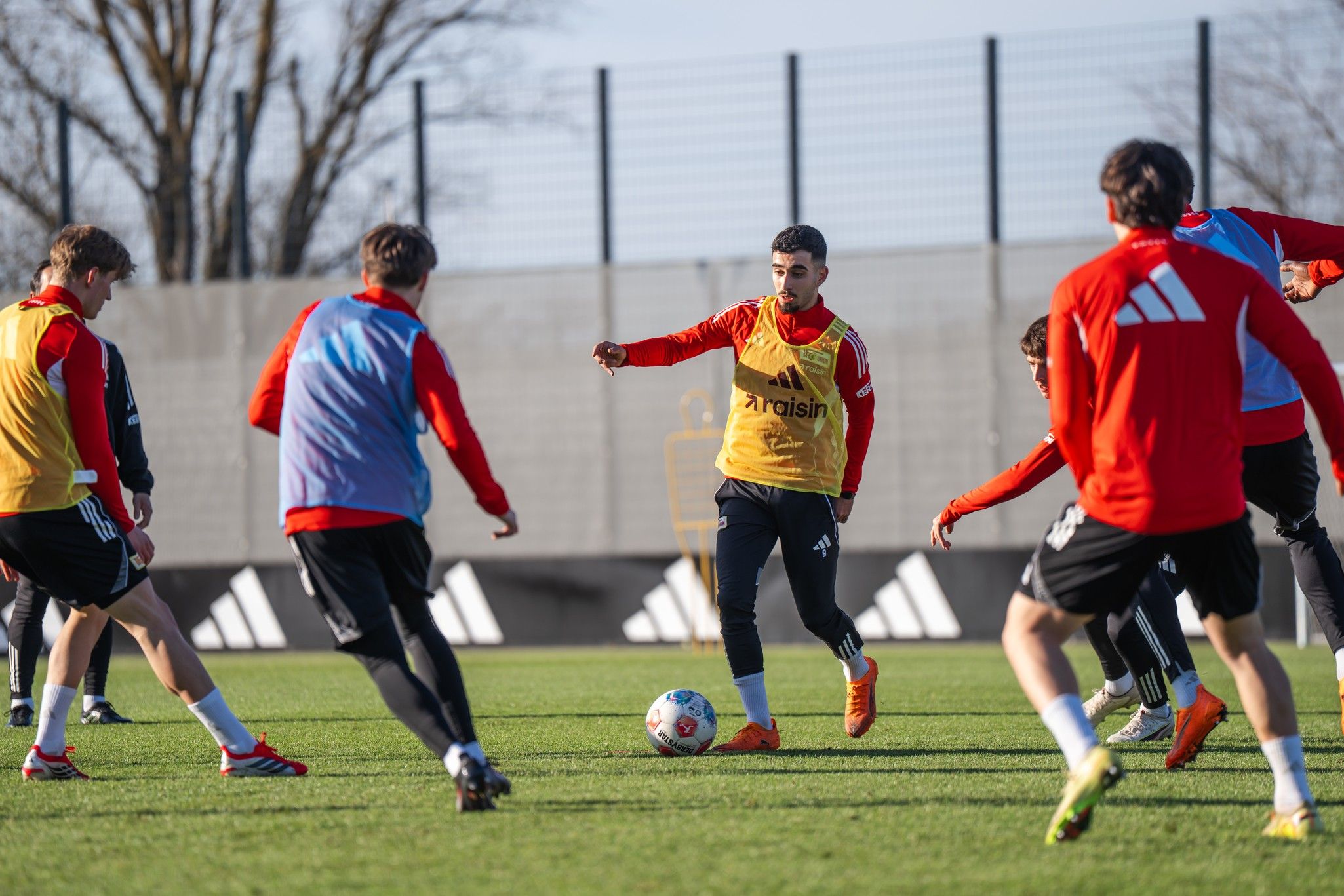 Ein Fußballtraining auf einem Rasenplatz mit mehreren Spielern in roten und blauen Trainingsanzügen, die den Ball umspielen.