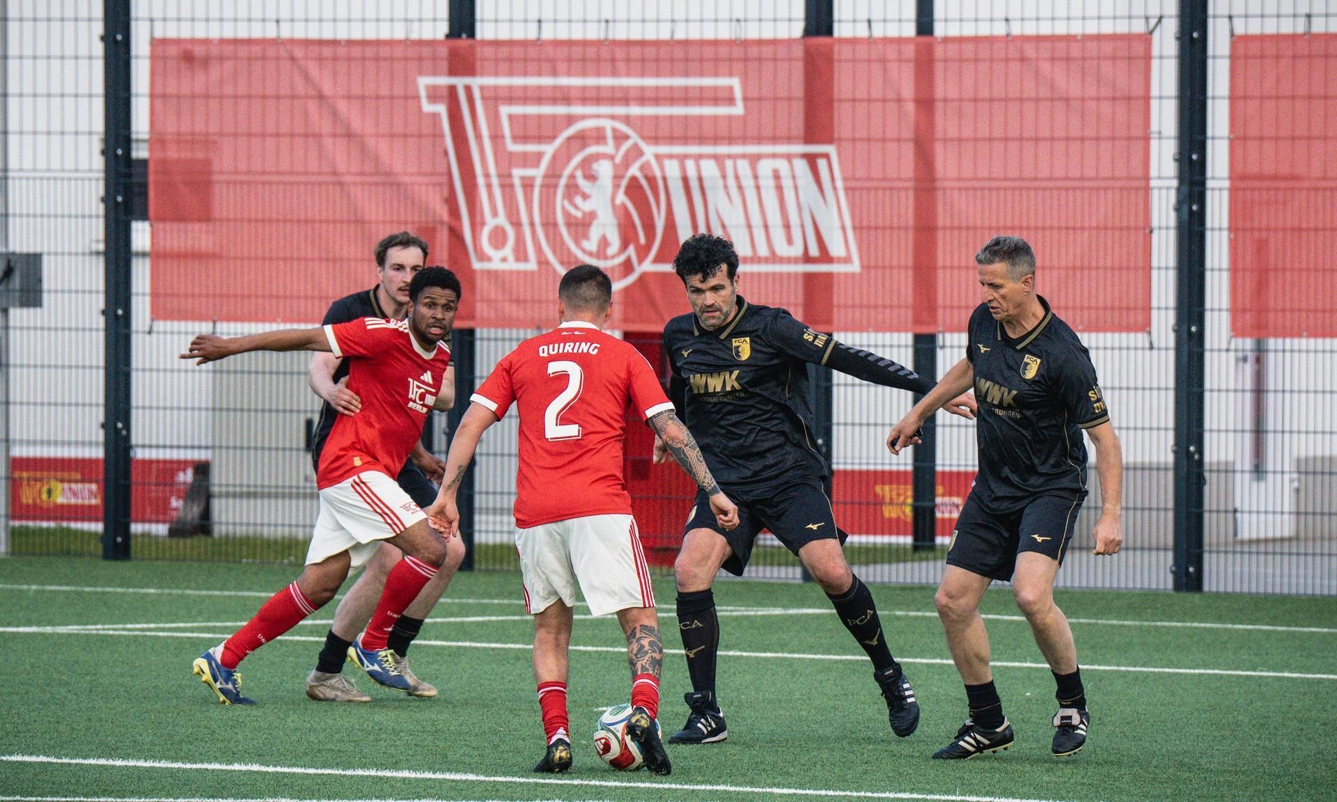 Fußballspiel mit Spielern in roten und schwarzen Trikots auf einem Kunstrasenfeld.
