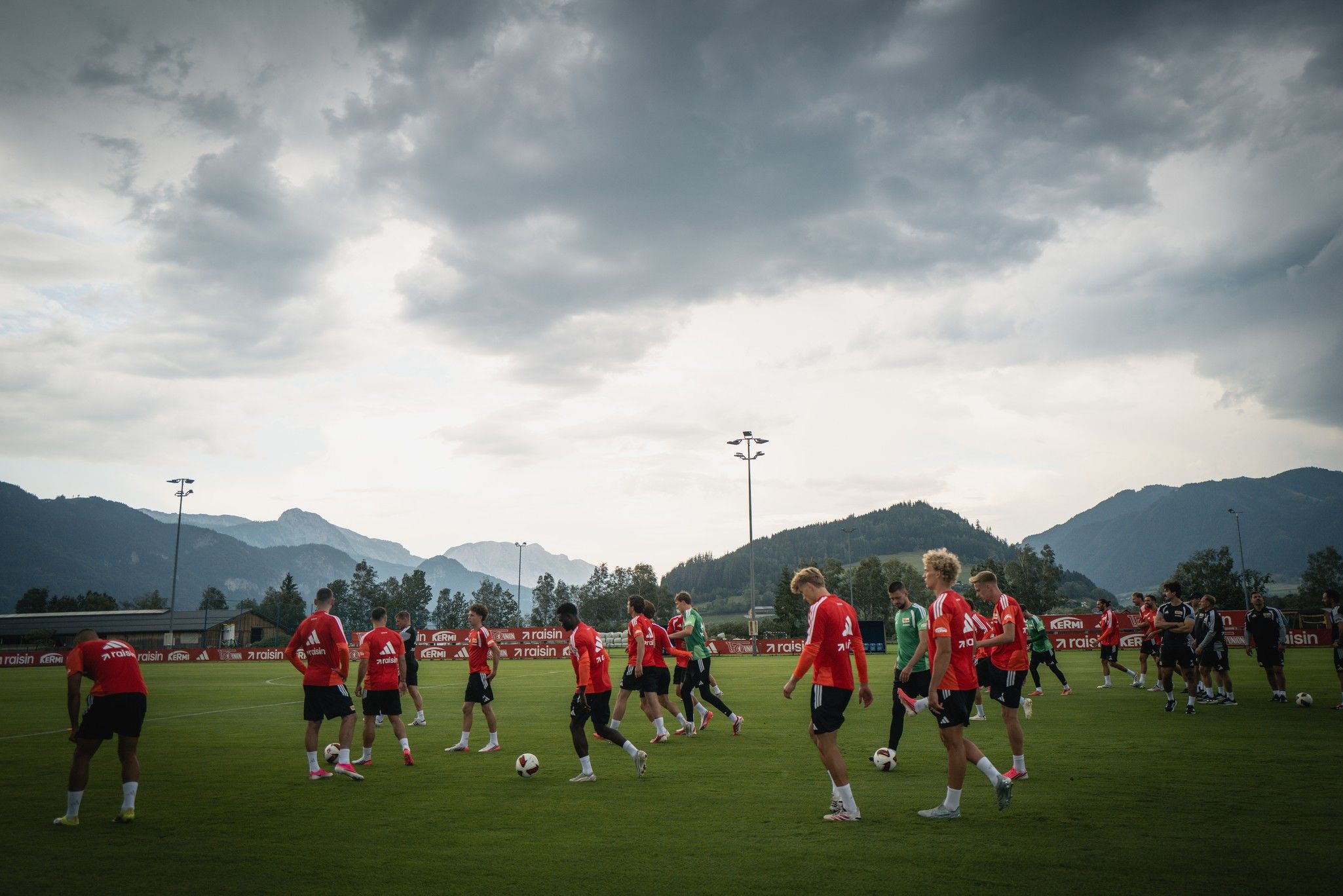 Fußballmannschaft in rot gekleideten Spielern trainiert auf einem Platz mit Bergen im Hintergrund unter bewölktem Himmel.