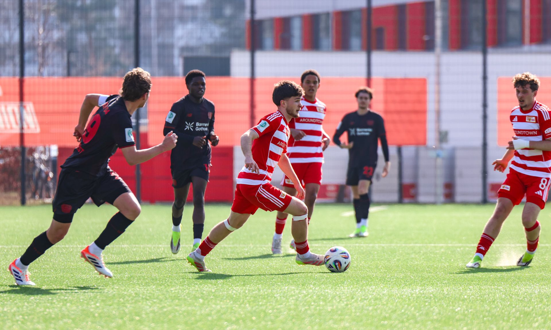 Junge Fußballspieler in rot-weißen Trikots dribbeln den Ball auf einem grünen Spielfeld, während Gegner versuchen, ihn zu verteidigen.