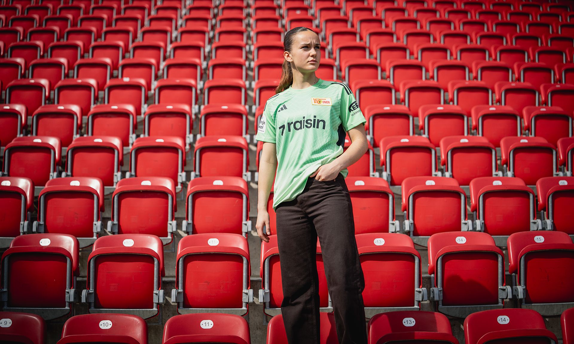 Eine Person in einem grünen Trikot steht inmitten leerer roter Stadionplätze.