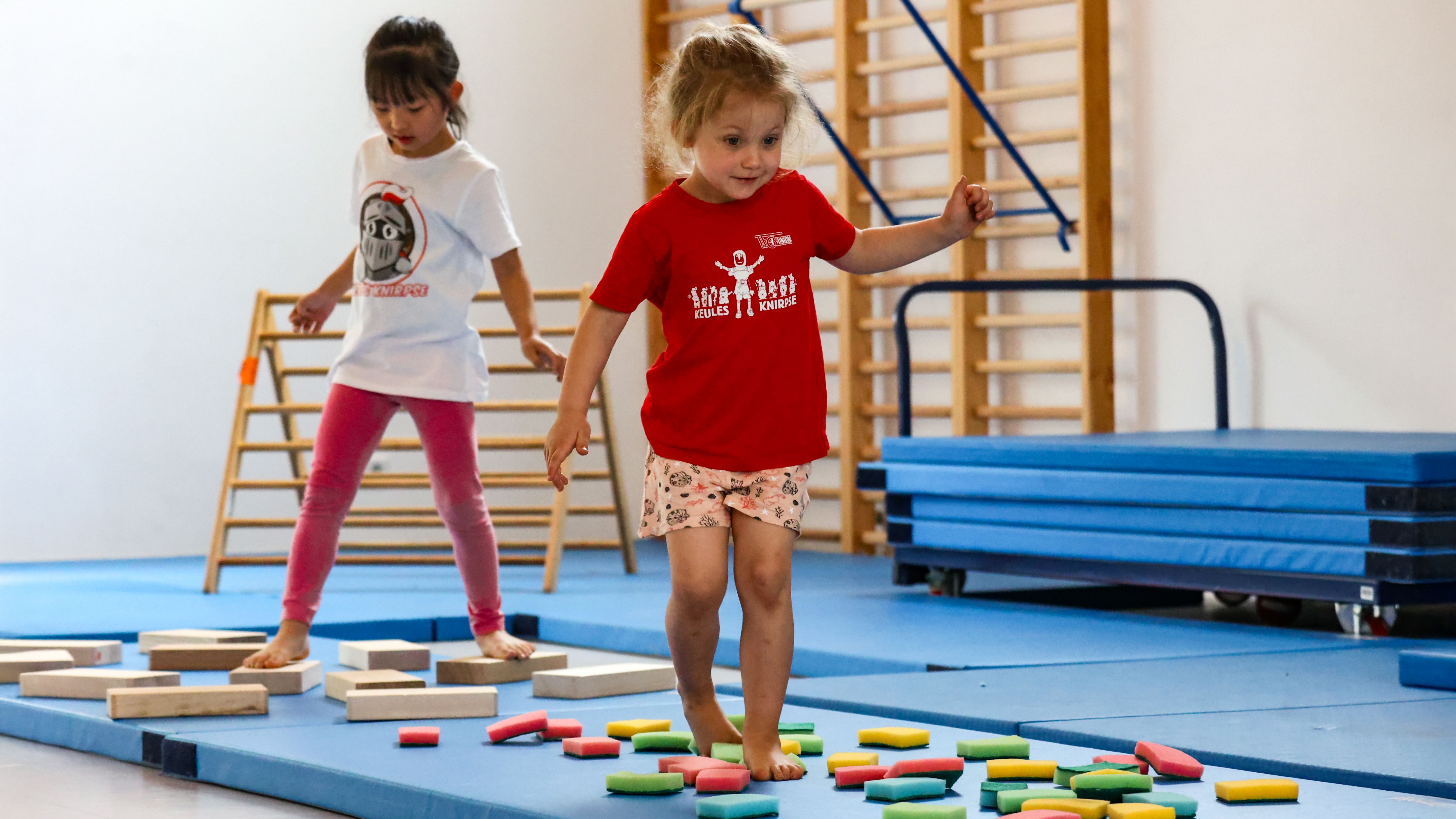 Ein kleines Mädchen in einem roten T-Shirt läuft über bunte, flache Steine auf einer Turnmatte, während ein anderes Mädchen im Hintergrund steht.