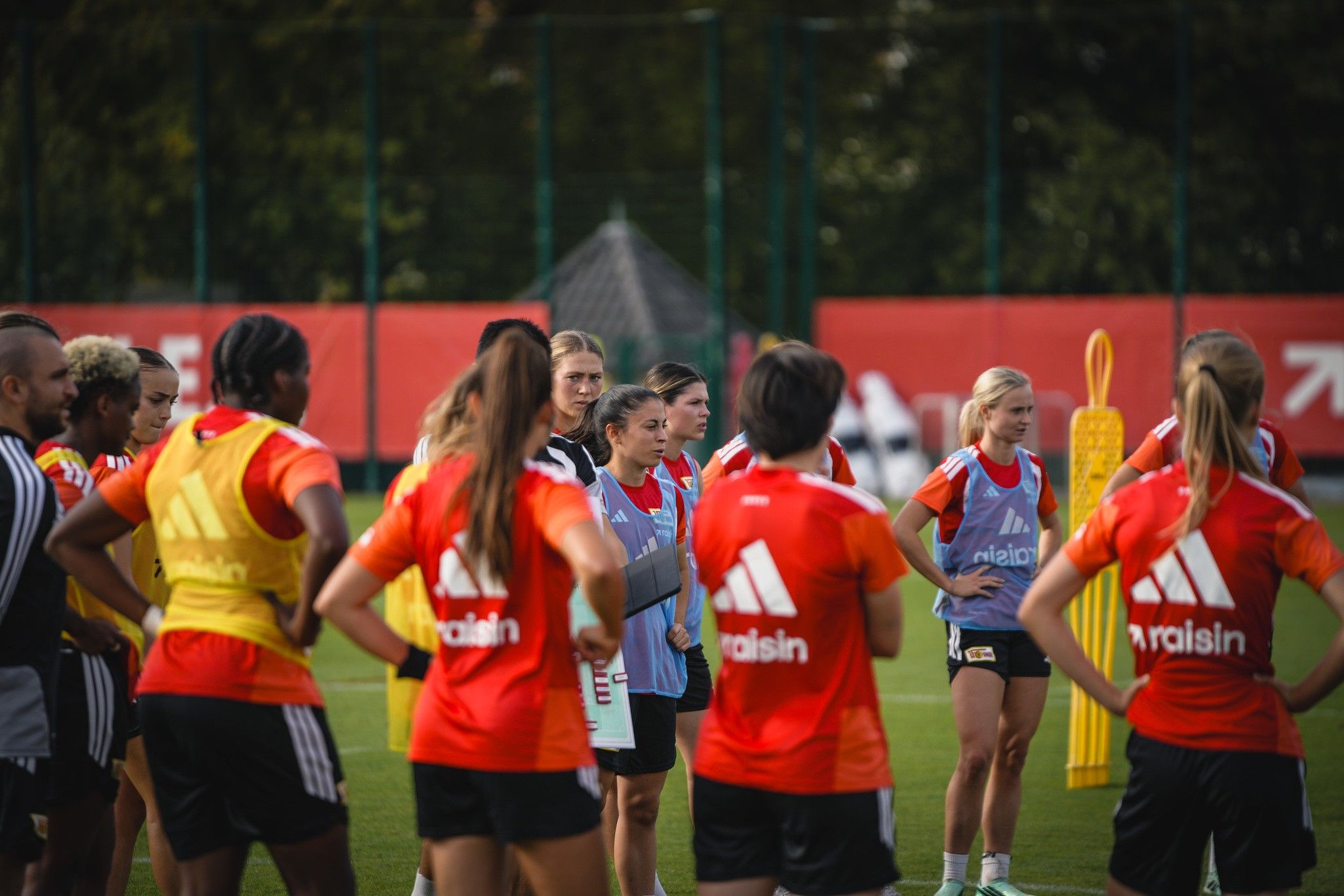 Frauenfußballmannschaft bei einem Training, Trainerin spricht mit den Spielerinnen, die in roten und blauen Trikots versammelt sind.