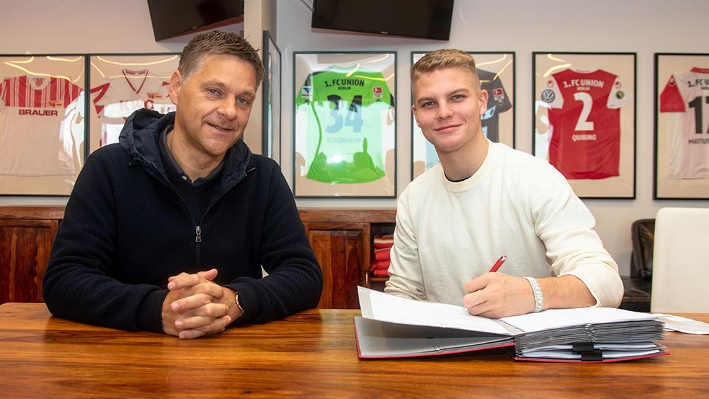 Zwei Männer sitzen an einem Tisch, der eine unterschreibt einen Vertrag, während der andere lächelt. Hinter ihnen hängen Fußballtrikots.