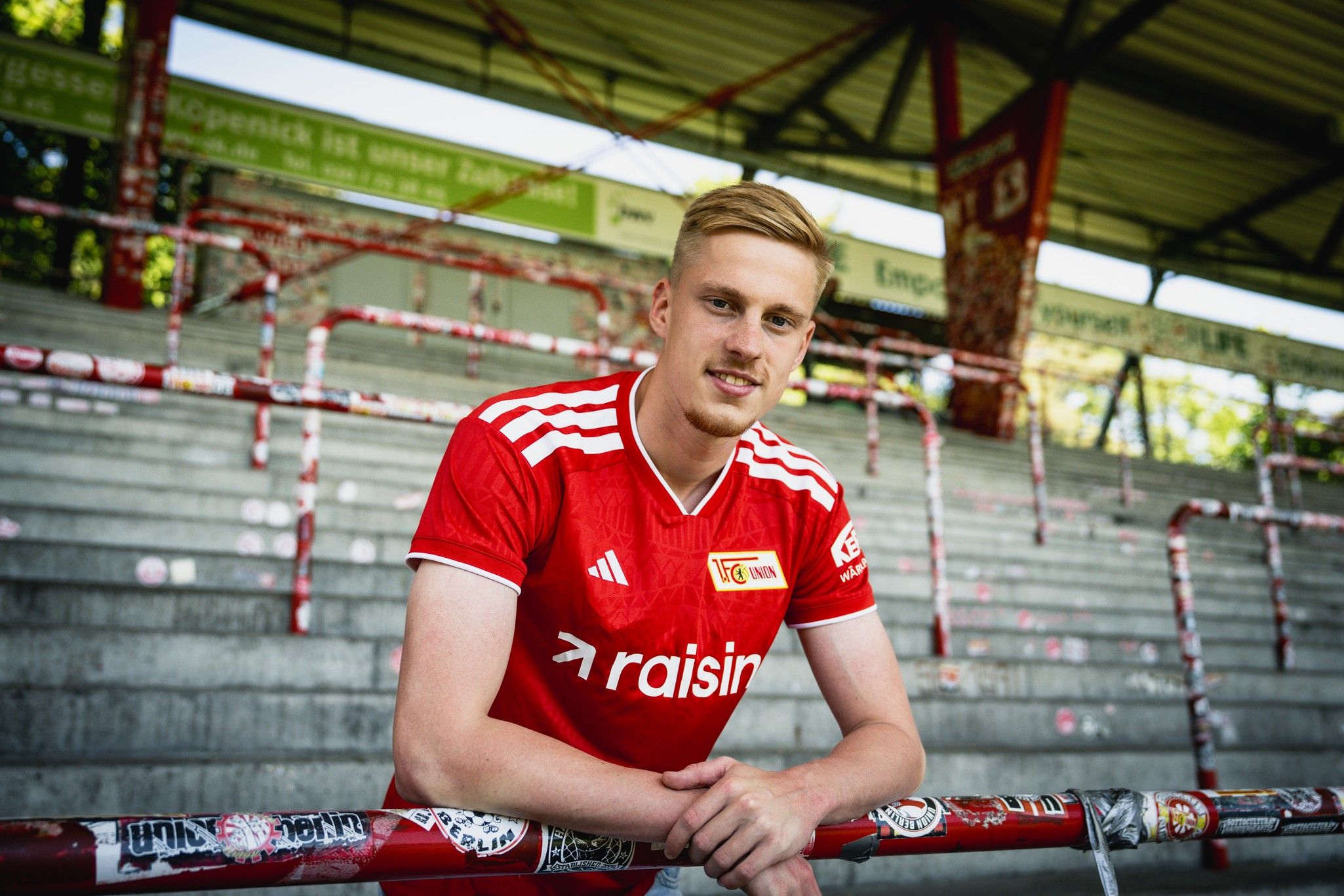 Ein junger Mann in einem roten Fußballtrikot steht lächelnd an einem Geländer im Stadion mit leeren Sitzplätzen im Hintergrund.