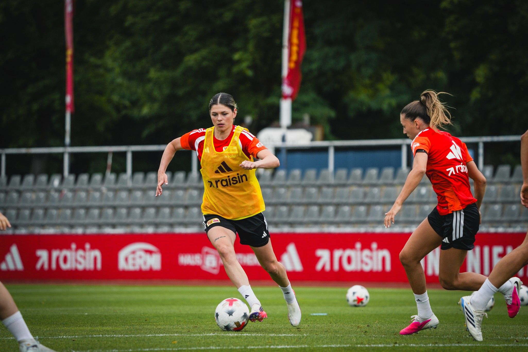 Eine weibliche Fußballspielerin in gelber Trikot läuft mit dem Ball, während zwei Teamkolleginnen im Hintergrund trainieren.