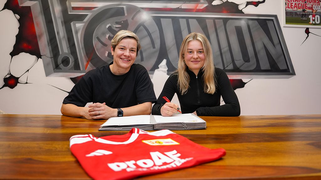 Zwei Frauen sitzen an einem Tisch, unterschreiben Verträge und posieren neben einem roten Fußballtrikot.
