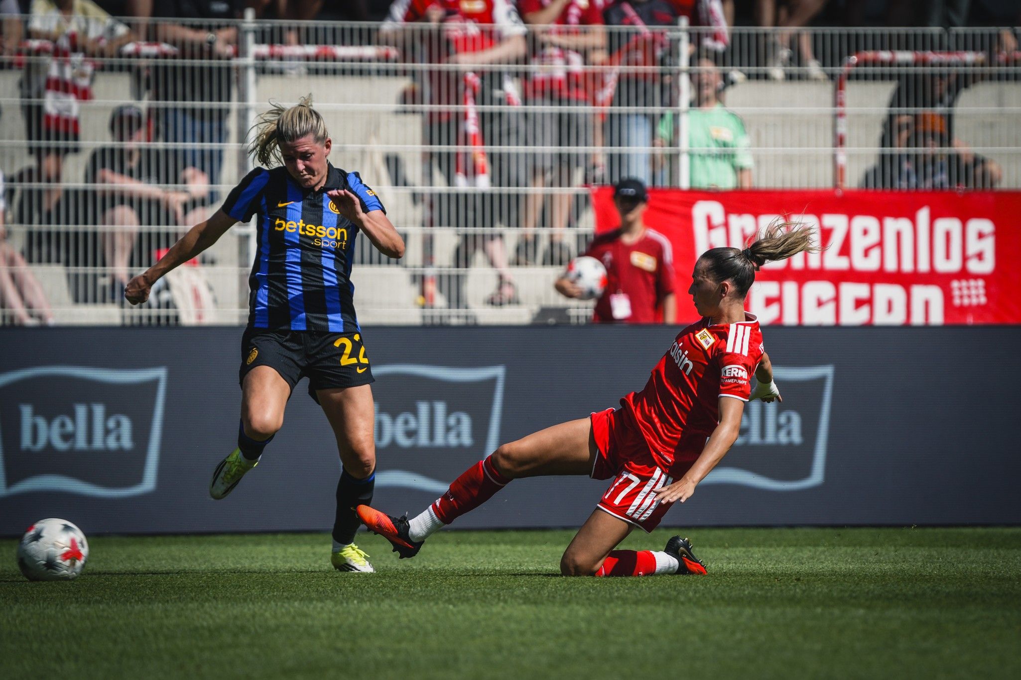 Eine Spielerin im blauen Trikot dribbelt, während eine andere im roten Trikot sie im Rasen foulspielt. Stadionatmosphäre im Hintergrund.