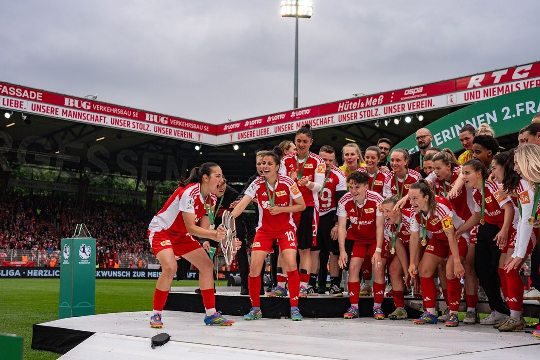 Frauenfußballmannschaft feiert den Gewinn eines Titels auf der Siegerbühne, umgeben von jubelnden Fans im Stadion.