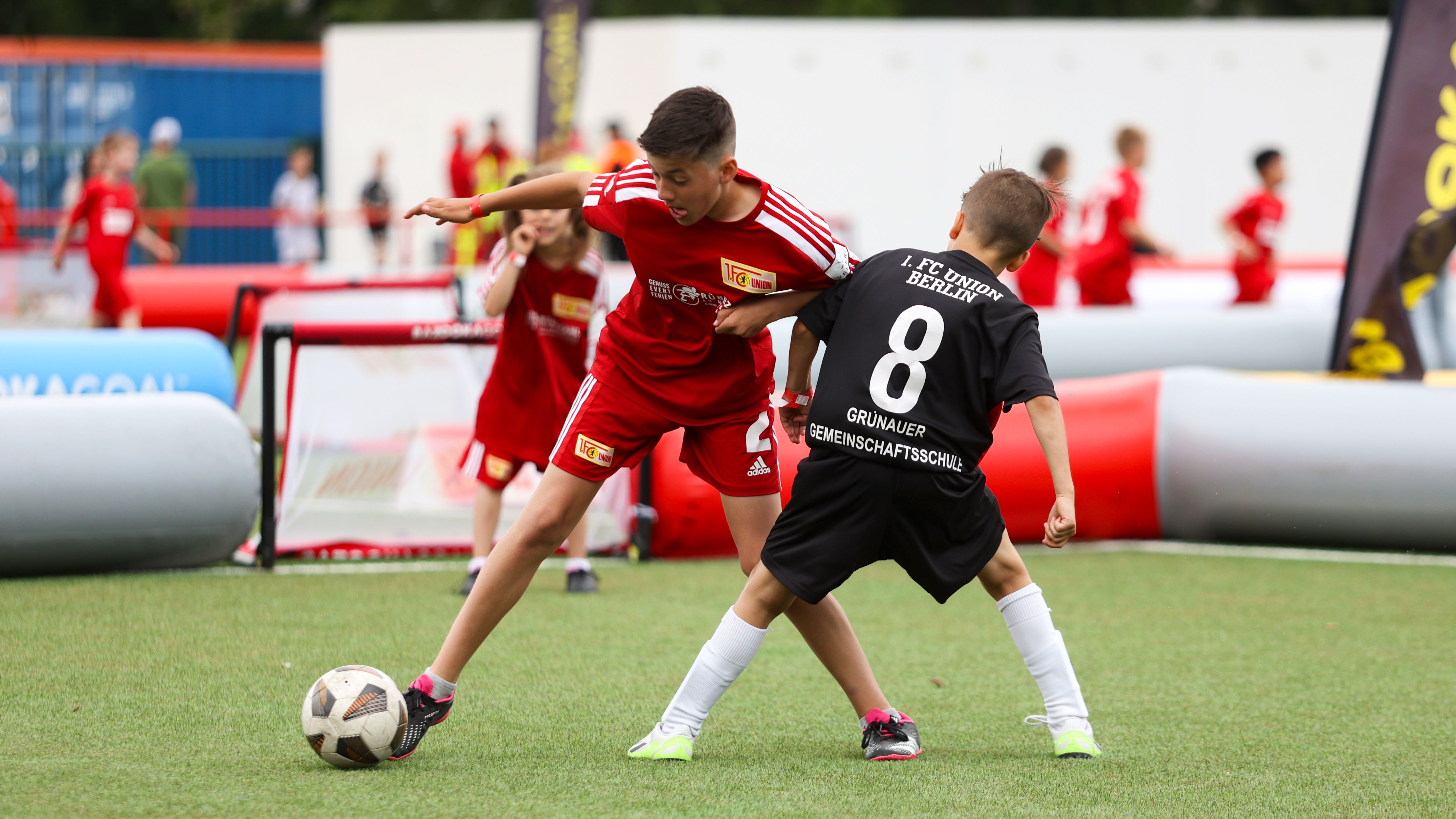 Zwei Jungen spielen Fußball auf einem grünen Platz, einer trägt ein rotes Trikot, der andere ein schwarzes.