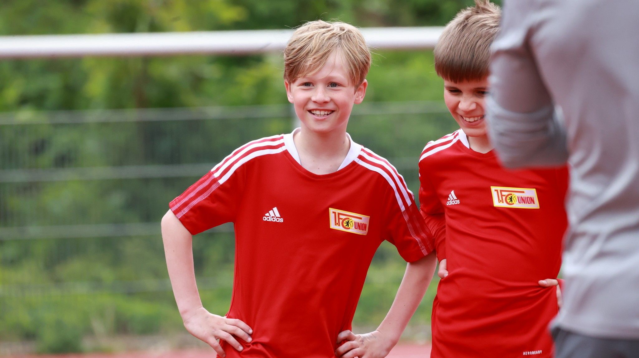 Zwei junge Fußballspieler in roten Trikots stehen lächelnd auf dem Spielfeld und posieren.