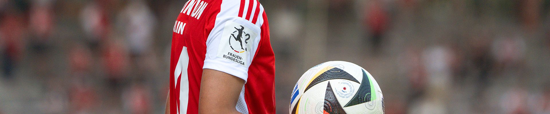 Ein Fußballspieler in rotem Trikot hält einen Fußball in der Hand, im Hintergrund sind Zuschauer auf den Rängen zu sehen.