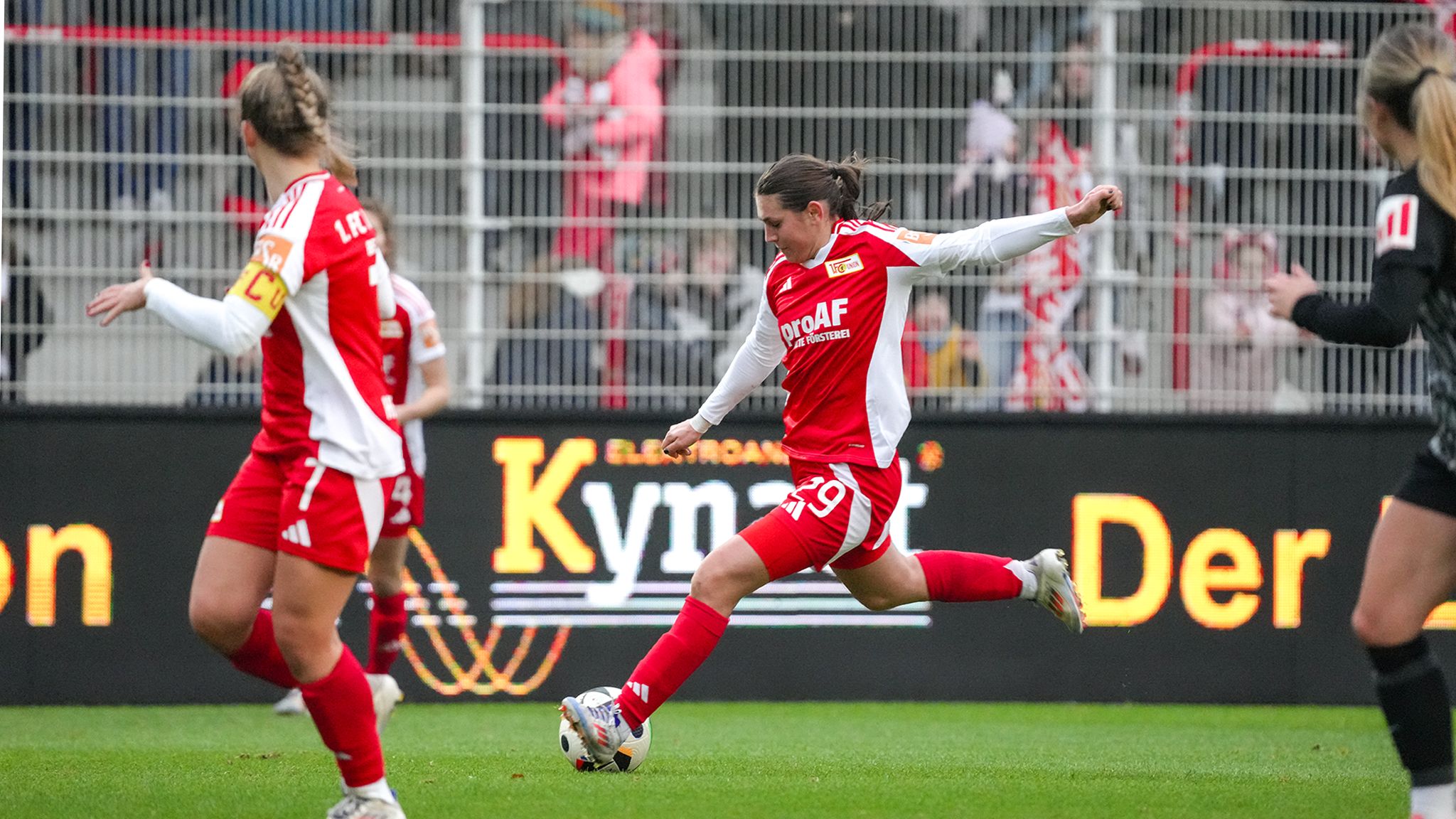 Eine Spielerin in rotem Trikot tritt den Ball während eines Fußballspiels, umgeben von anderen Teamkolleginnen.