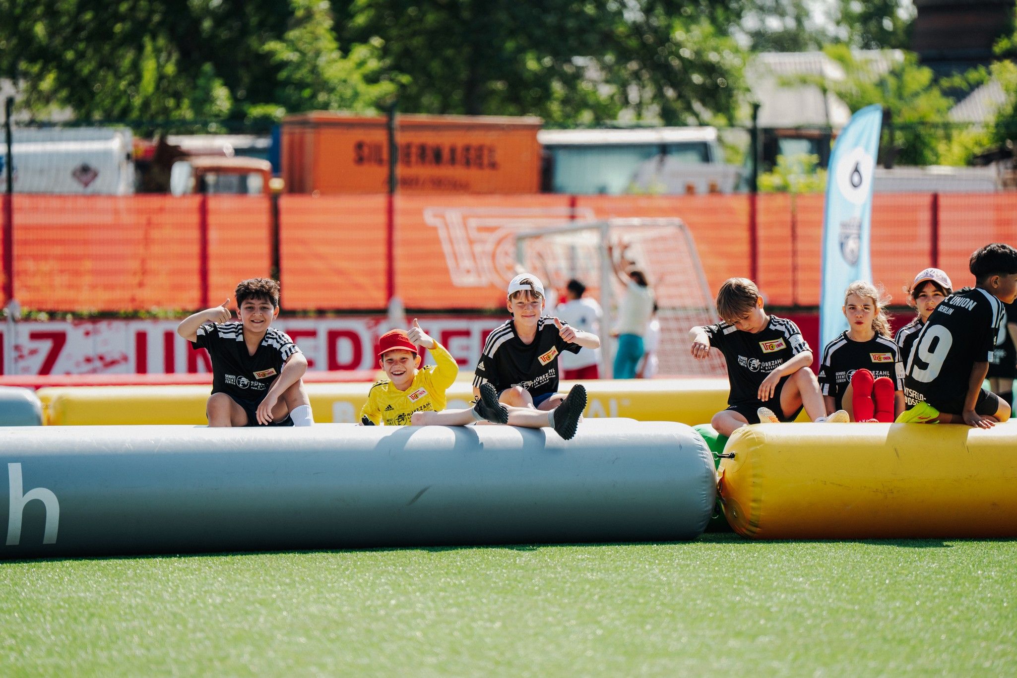 Kinder in Sportklamotten sitzen auf großen Schaumstoffkissen und lächeln. Es ist ein sonniger Tag auf einem Sportgelände.