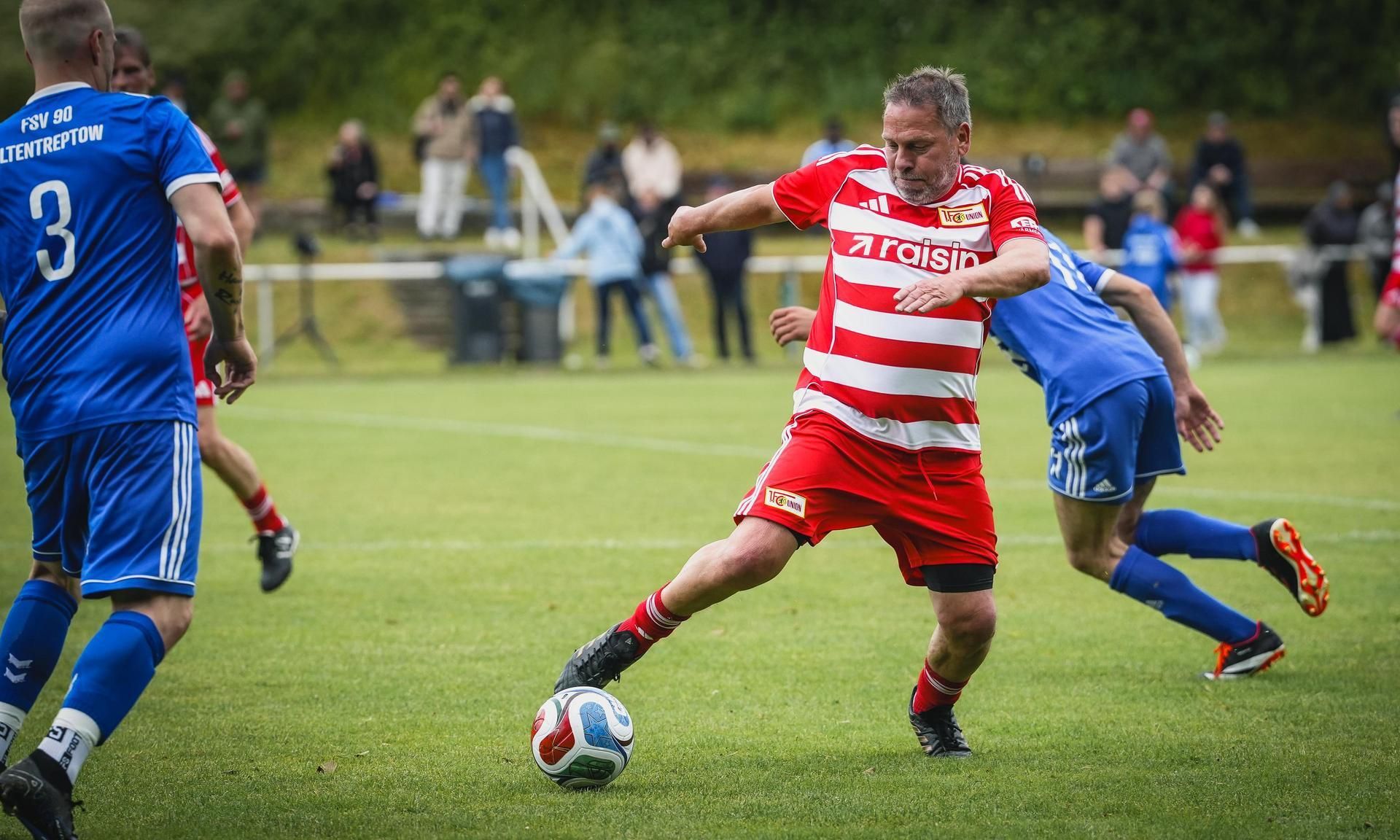 Ein Spieler in rot-weißer Uniform tritt den Ball auf einem Fußballplatz, während ein Gegner in blauer Uniform ihn verfolgt.