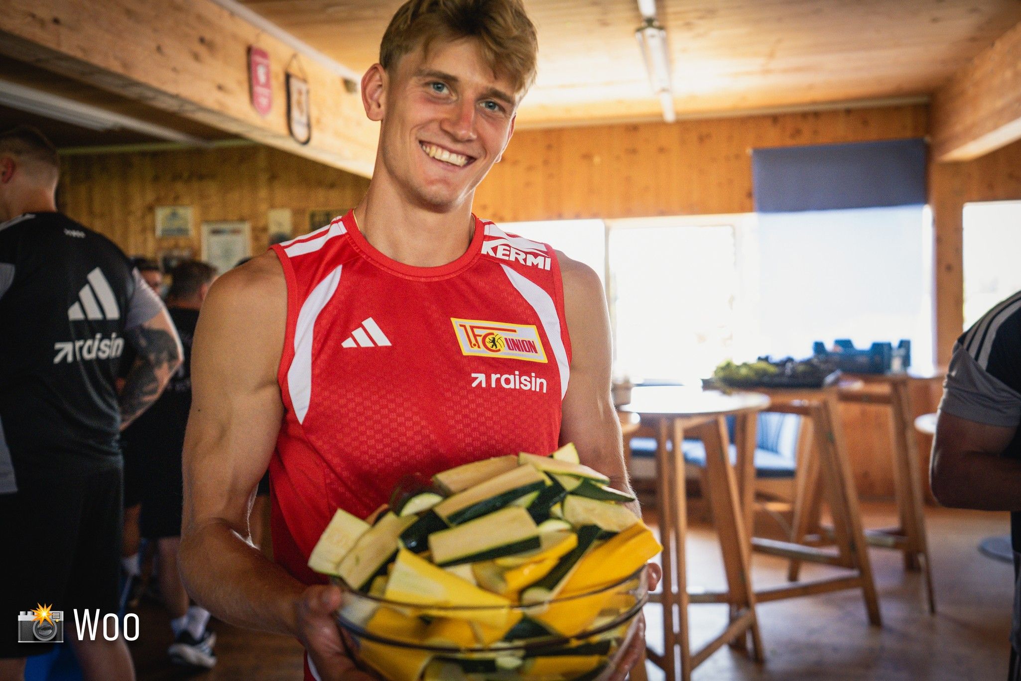 Ein junger Mann in einem roten Trikot hält eine große Schüssel mit verschiedenen Gemüsesorten in einer Holzhalle.