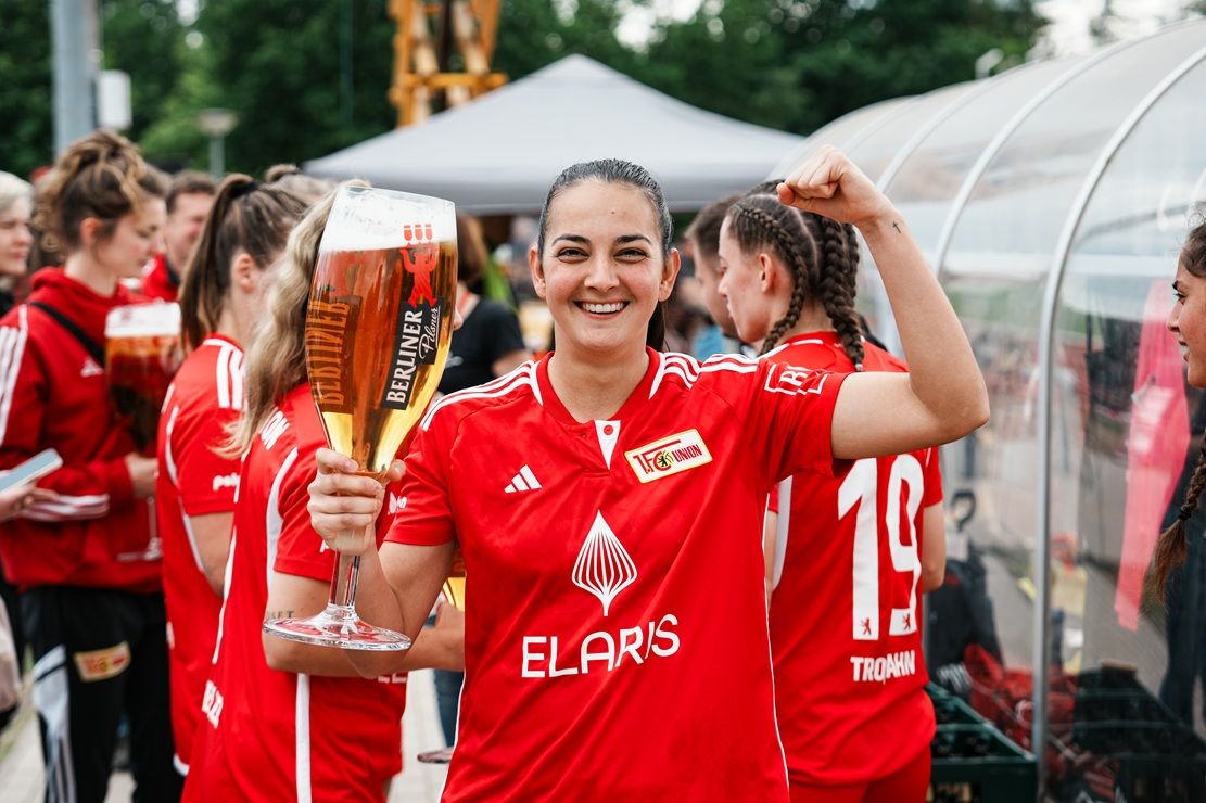 Eine lächelnde Frau in einem roten Trikot hebt ein großes Bierglas und zeigt den Siegessymbol mit einer Hand. Im Hintergrund Gruß von anderen.