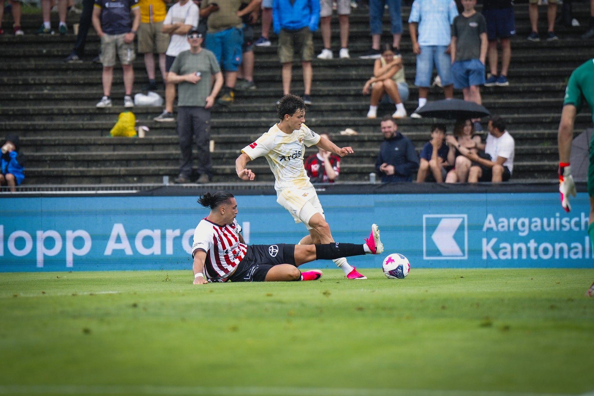 Ein Fußballspieler umgeht einen Gegner, während Zuschauer auf der Tribüne zuschauen.