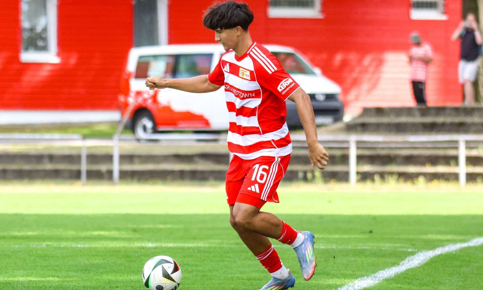 Ein Fußballspieler in rotem Trikot mit weißen Streifen führt einen Ball auf einem grünen Feld.
