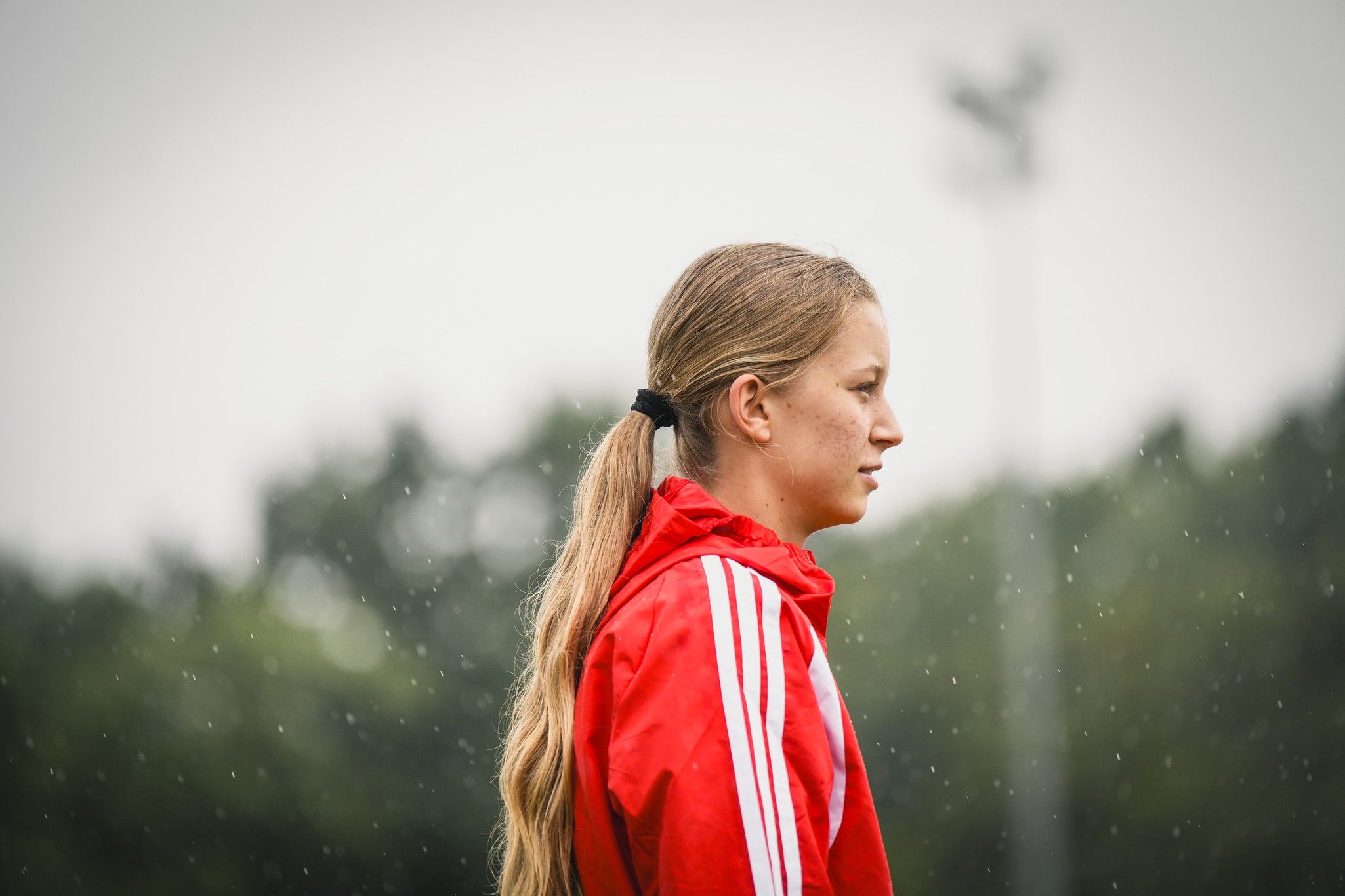 Eine sportliche Person mit langem, blondem Haar in einem roten Trainingsanzug steht bei regnerischem Wetter im Profil.
