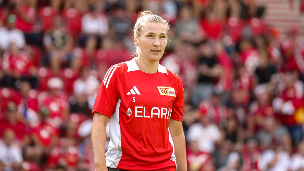 A player in a red jersey with the number 5 stands on the field, with many fans visible in the background.