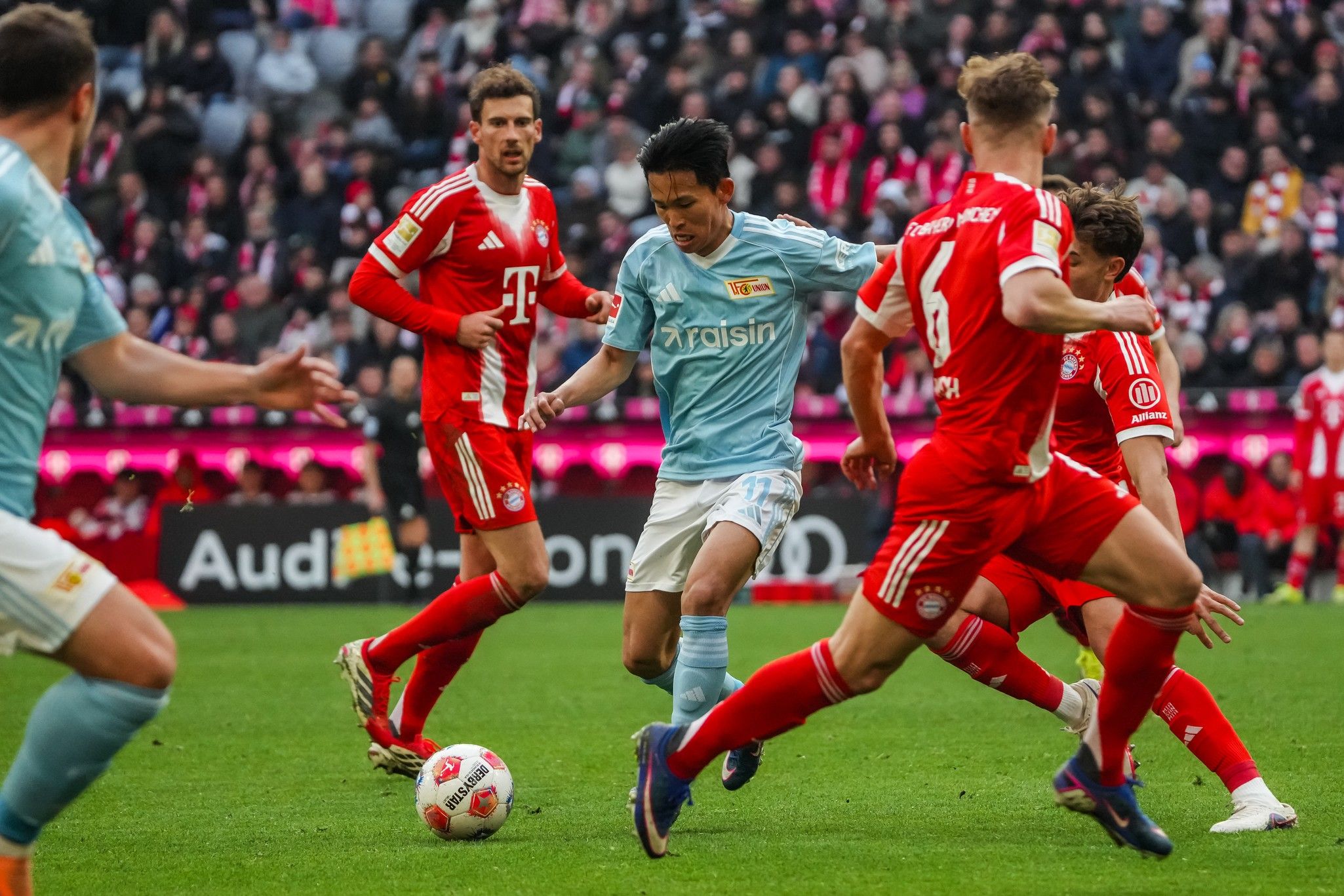 Spielszene eines Fußballspiels, in dem ein Spieler in blauer Kleidung von mehreren Spielern in Rot umgeben ist.