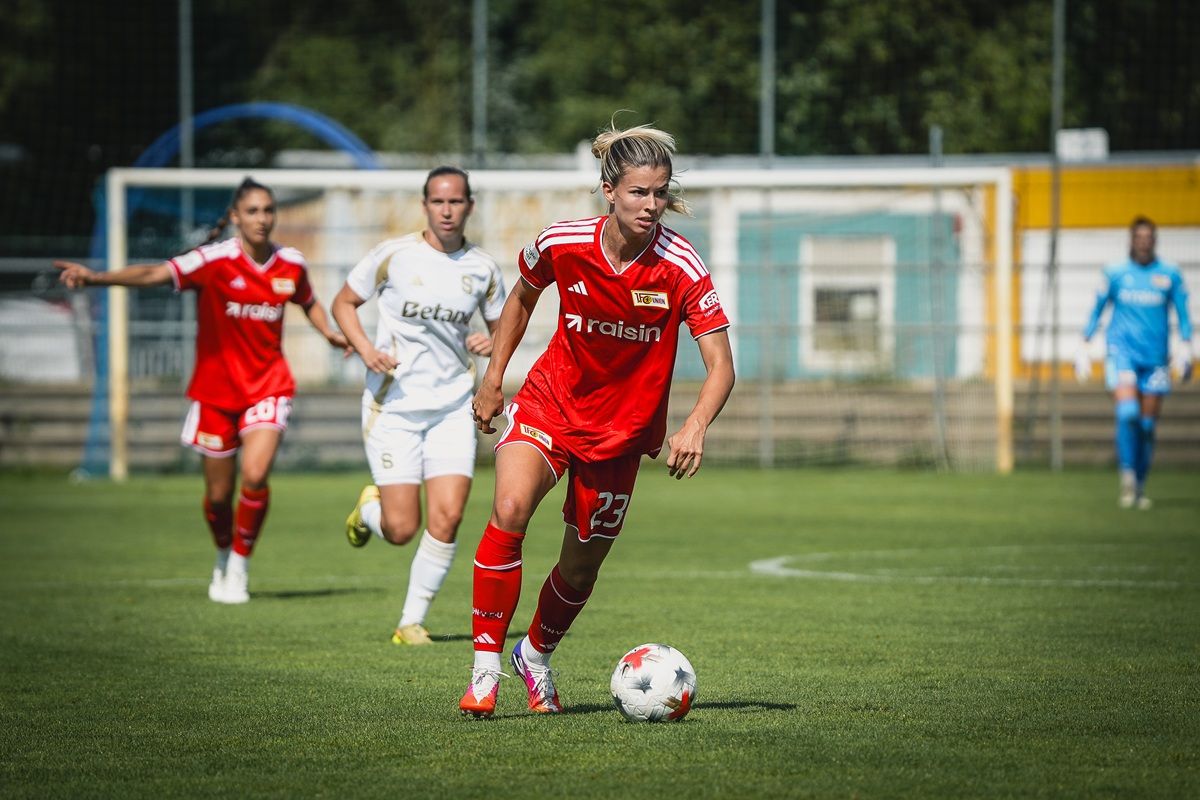 Eine Fußballspielerin in rotem Trikot dribbelt den Ball auf einem Sportplatz, während zwei andere Spielerinnen im Hintergrund stehen.