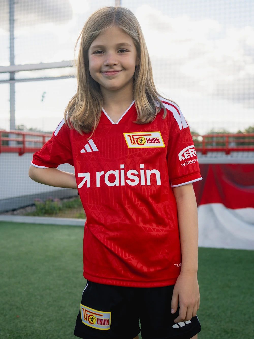 Mädchen mit langen Haaren trägt ein rotes Fußballtrikot mit dem Logo des 1. FC Union Berlin auf einem Sportplatz.