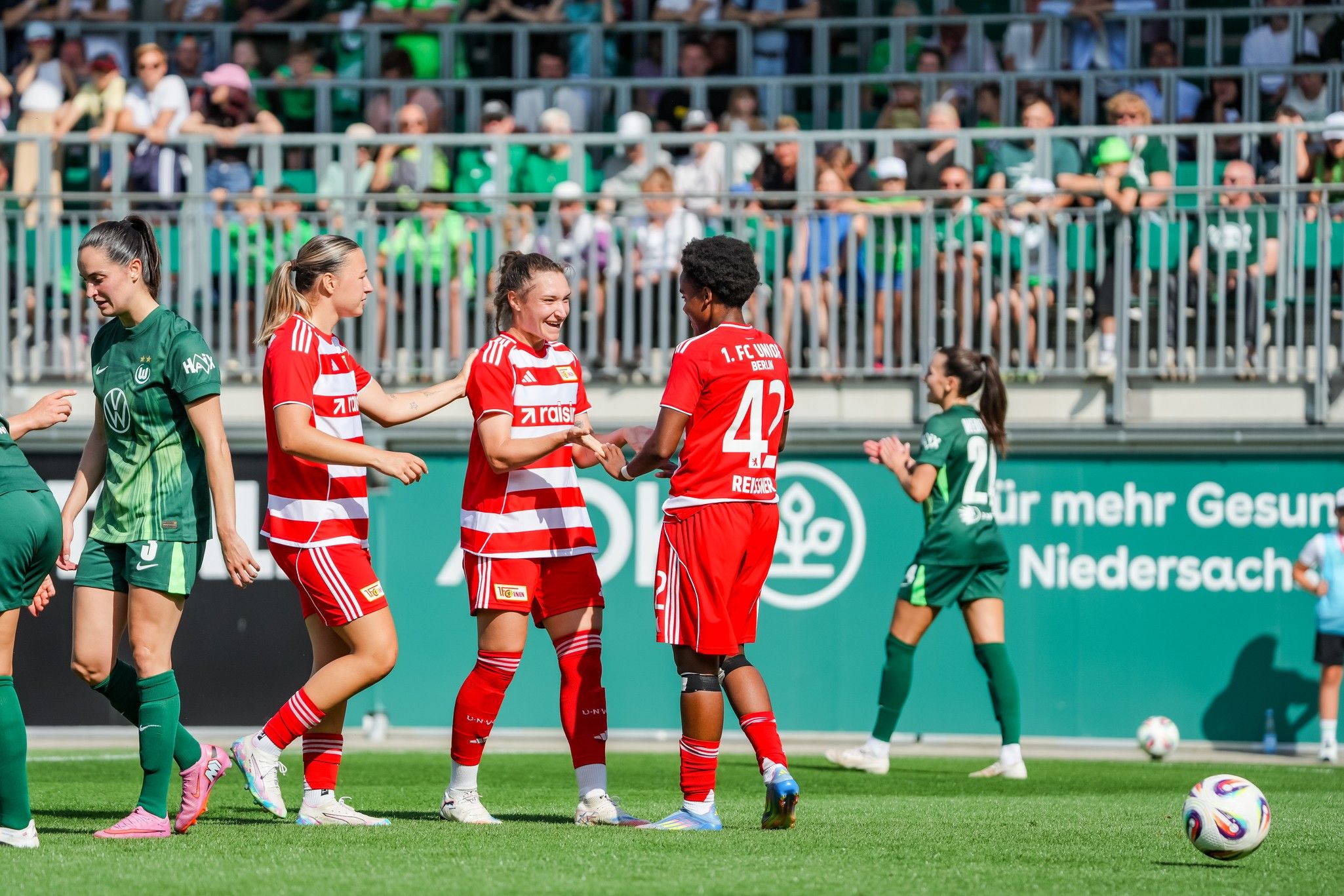 Drei Fußballspielerinnen in roten Trikots feiern einen Punktgewinn auf dem Platz, während andere Spielerinnen im Hintergrund positionsbedingt agieren.