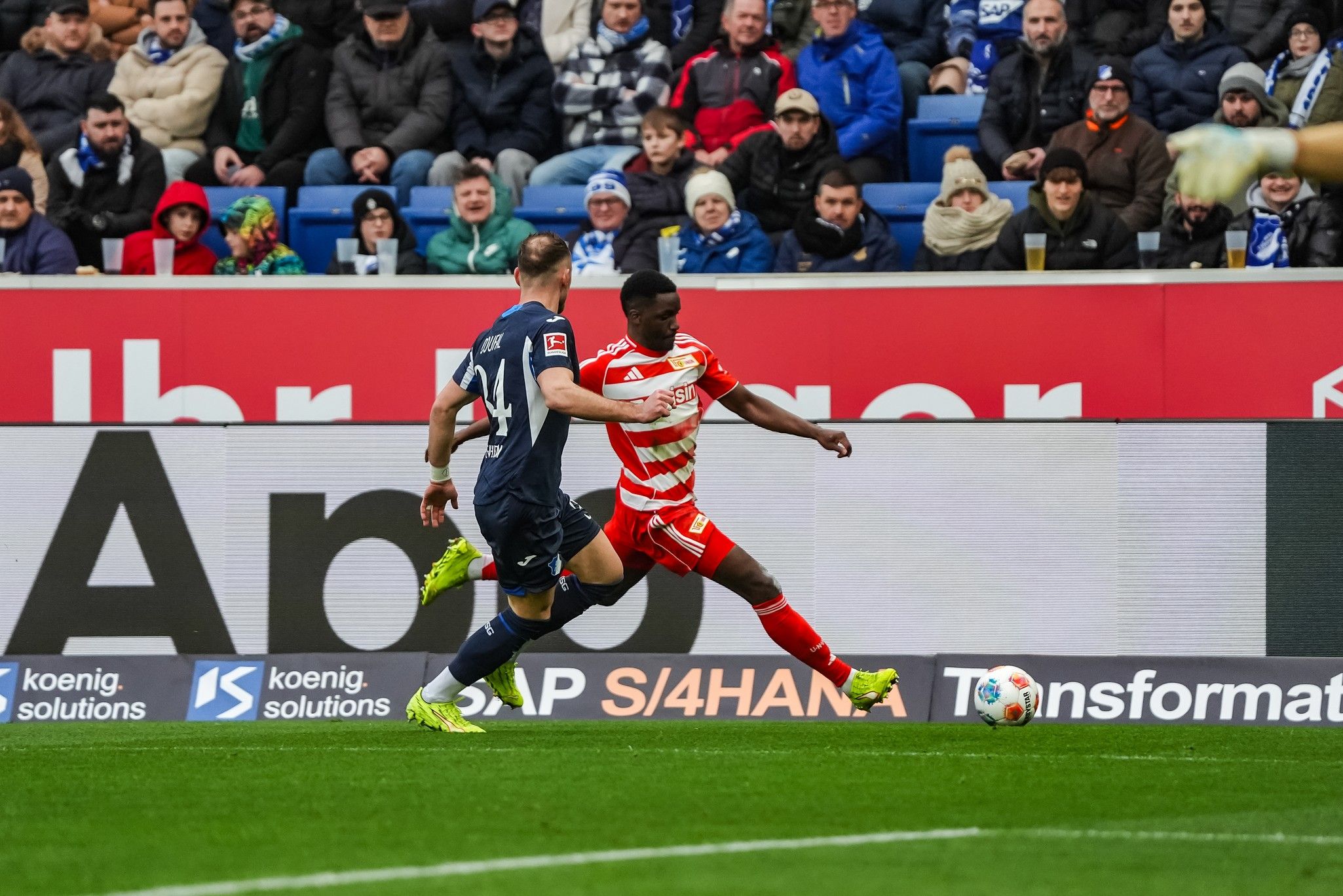 Ein Fußballspieler in rot-weißer Uniform dribbelt am Gegner in blauer Uniform vorbei, während Zuschauer im Hintergrund zuschauen.