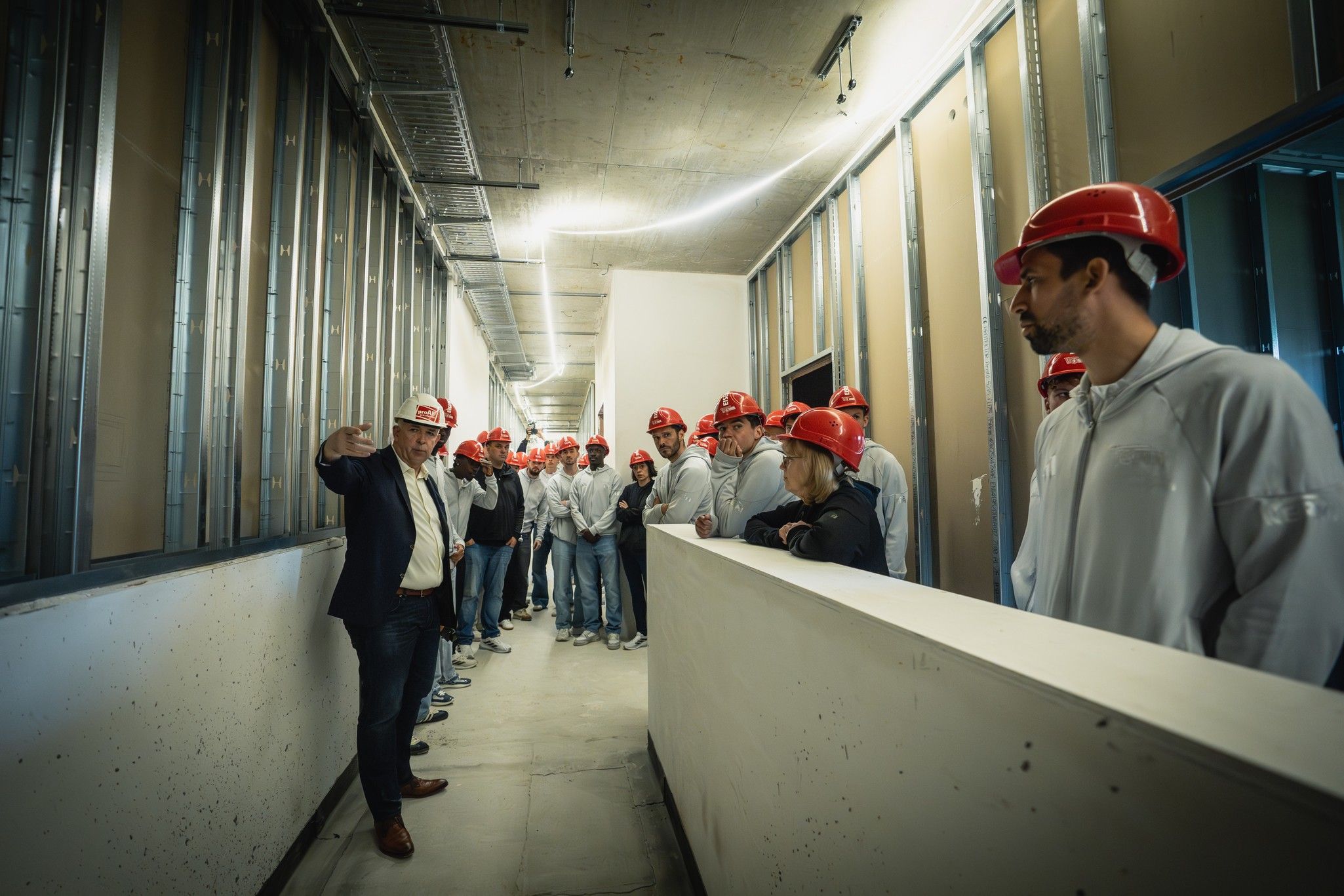 Gruppe von Bauarbeitern mit roten Helmen in einem Flur, während ein Mann mit Anzug eine Präsentation hält.