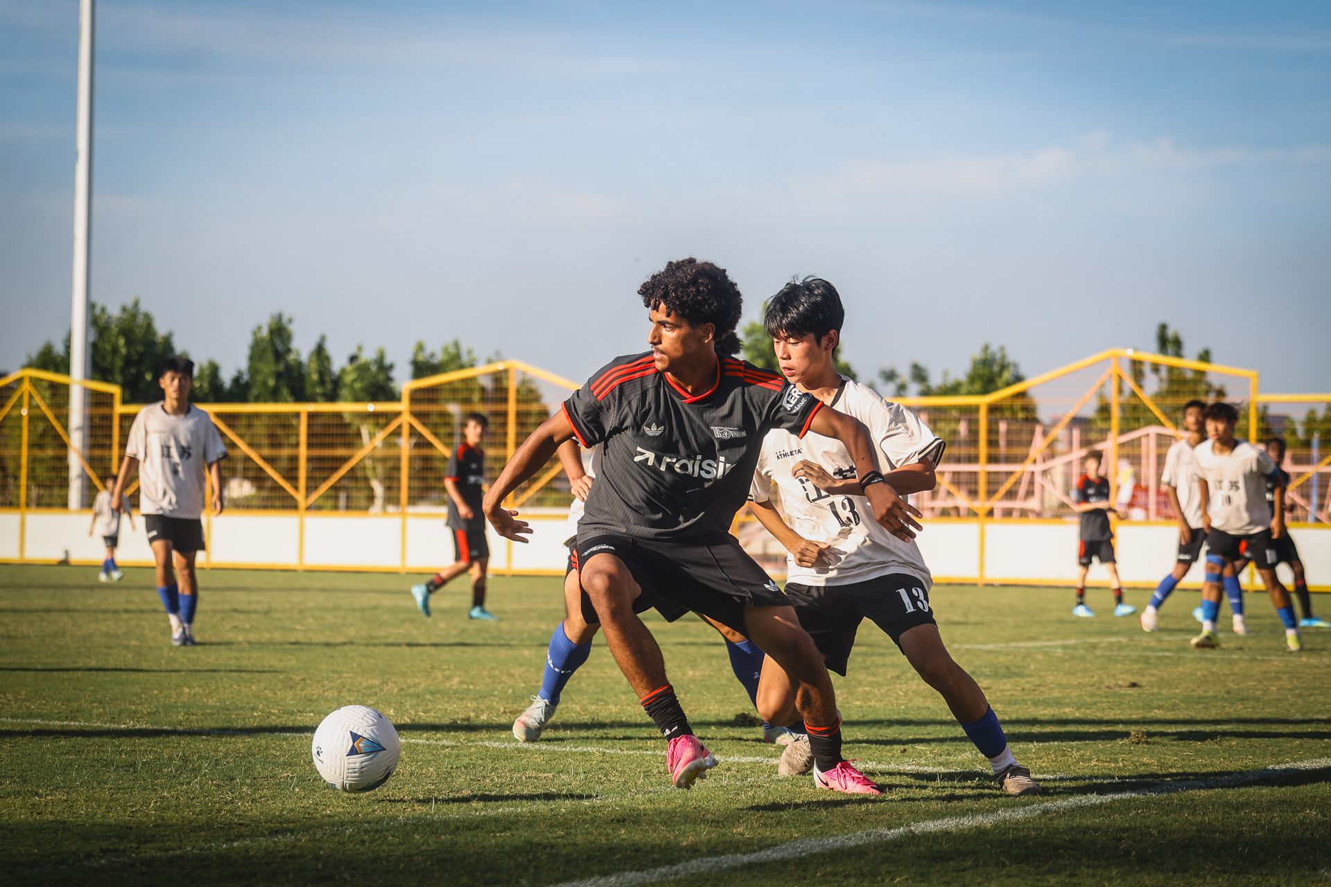 Zwei Jugendliche kämpfen um den Ball während eines Fußballspiels auf einem grünen Feld unter blauem Himmel.