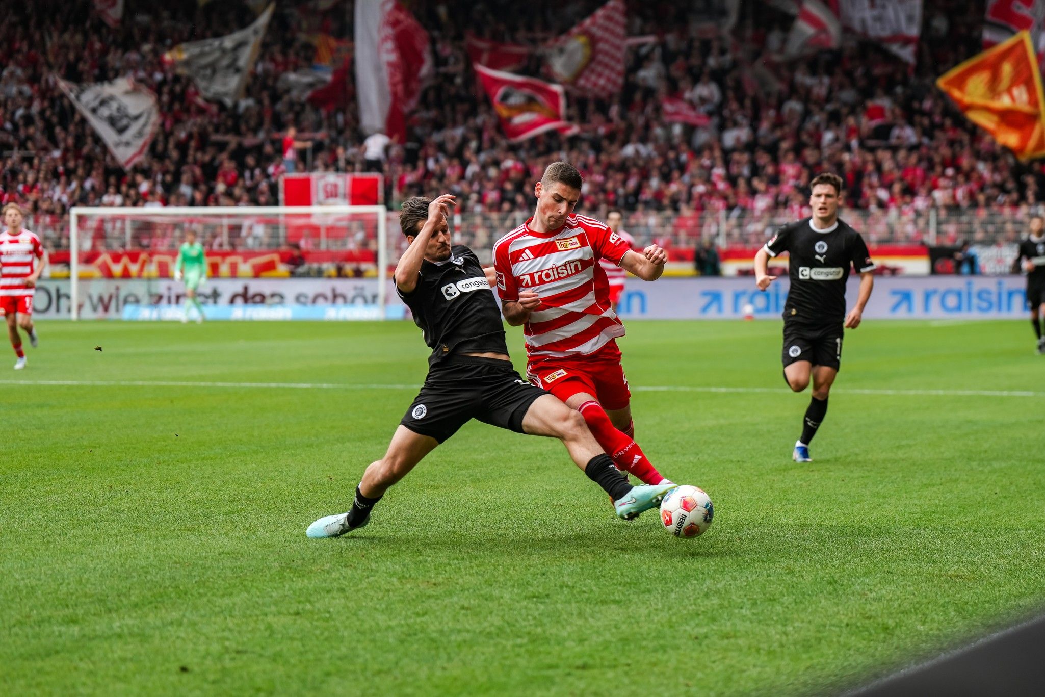 Zwei Fußballspieler kämpfen um den Ball im Stadion, umgeben von jubelnden Fans und Vereinsflaggen.