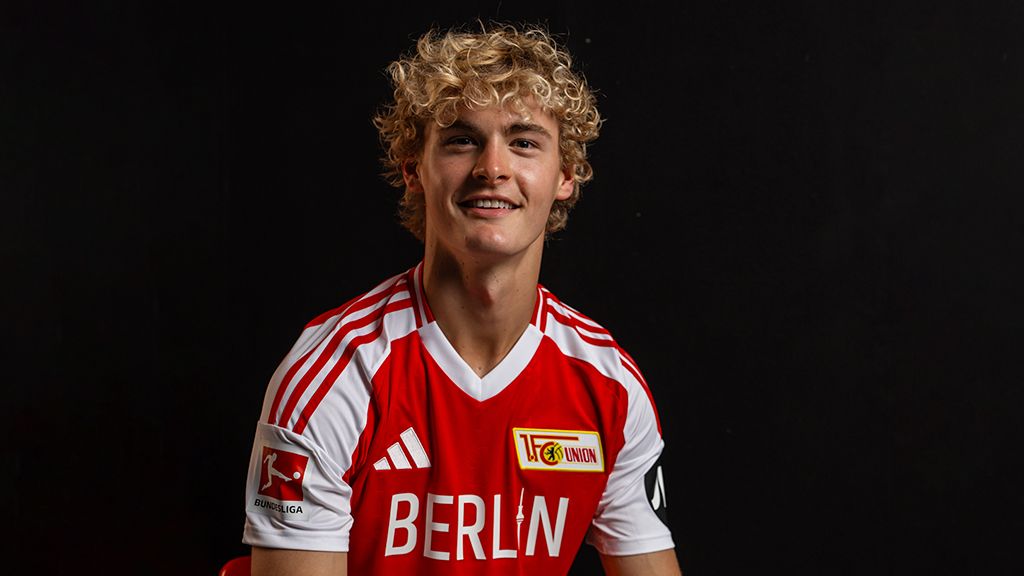 A young man with curly hair is wearing a red jersey with a football club logo. He is smiling at the camera.