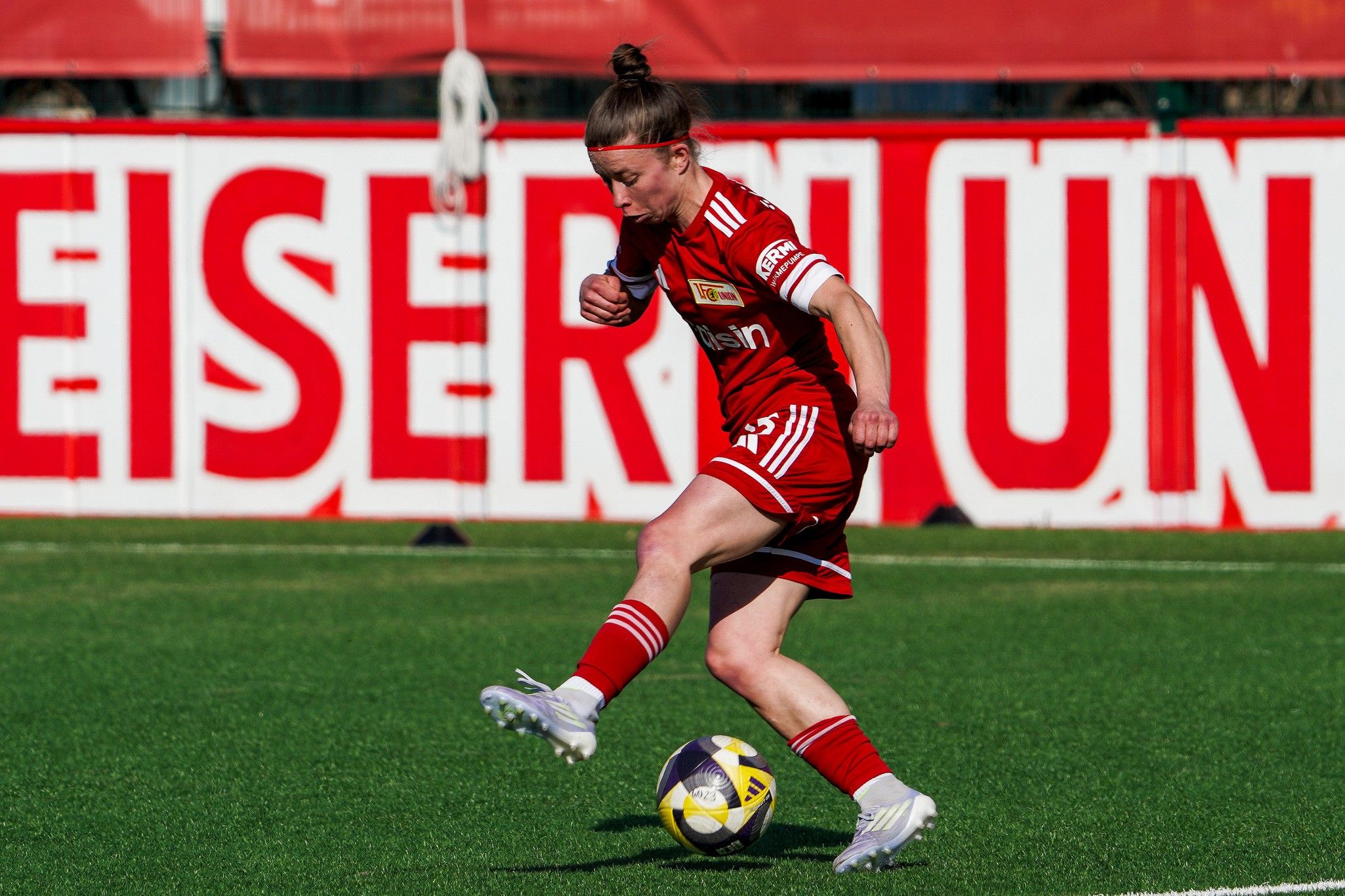 Eine Spielerin in rotem Trikot schießt den Ball auf einem Fußballfeld mit einem großen Banner im Hintergrund.