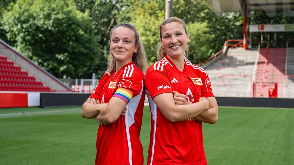 Zwei Frauen im roten Fußballtrikot stehen mit verschränkten Armen auf einem Stadionrasen.